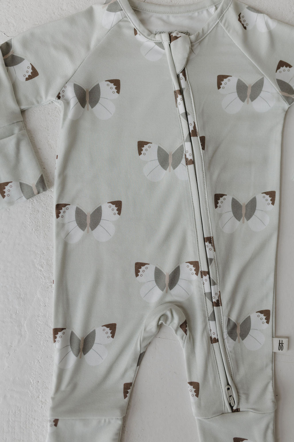 The forever french baby Bamboo Zip Pajamas in Butterflies feature a light gray design with brown and gray butterfly patterns. These hypoallergenic, long-sleeve pajamas have a front zipper from neck to leg and are crafted from soft, breathable fabric for comfort.