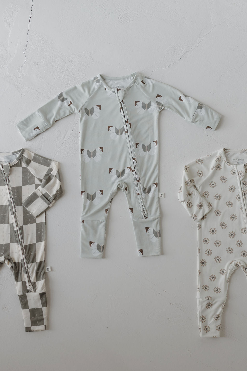 A trio of forever french baby Bamboo Zip Pajamas | Oat & Sage is displayed: gray with a white checkered pattern on the left, light blue with abstract shapes in the center, and cream with a small floral design on the right. Each hypo-allergenic piece includes a handy front zipper.