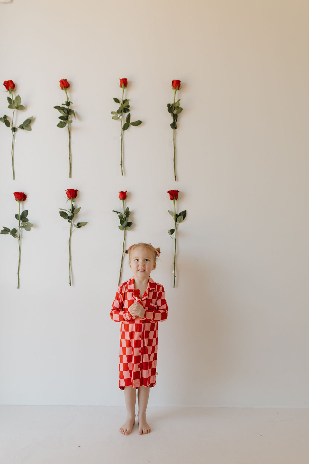 A young girl stands barefoot against a white wall in a Toddler Bamboo Sleeping Dress by forever french baby, holding a rose. Behind her, eight red roses are displayed vertically in two rows.