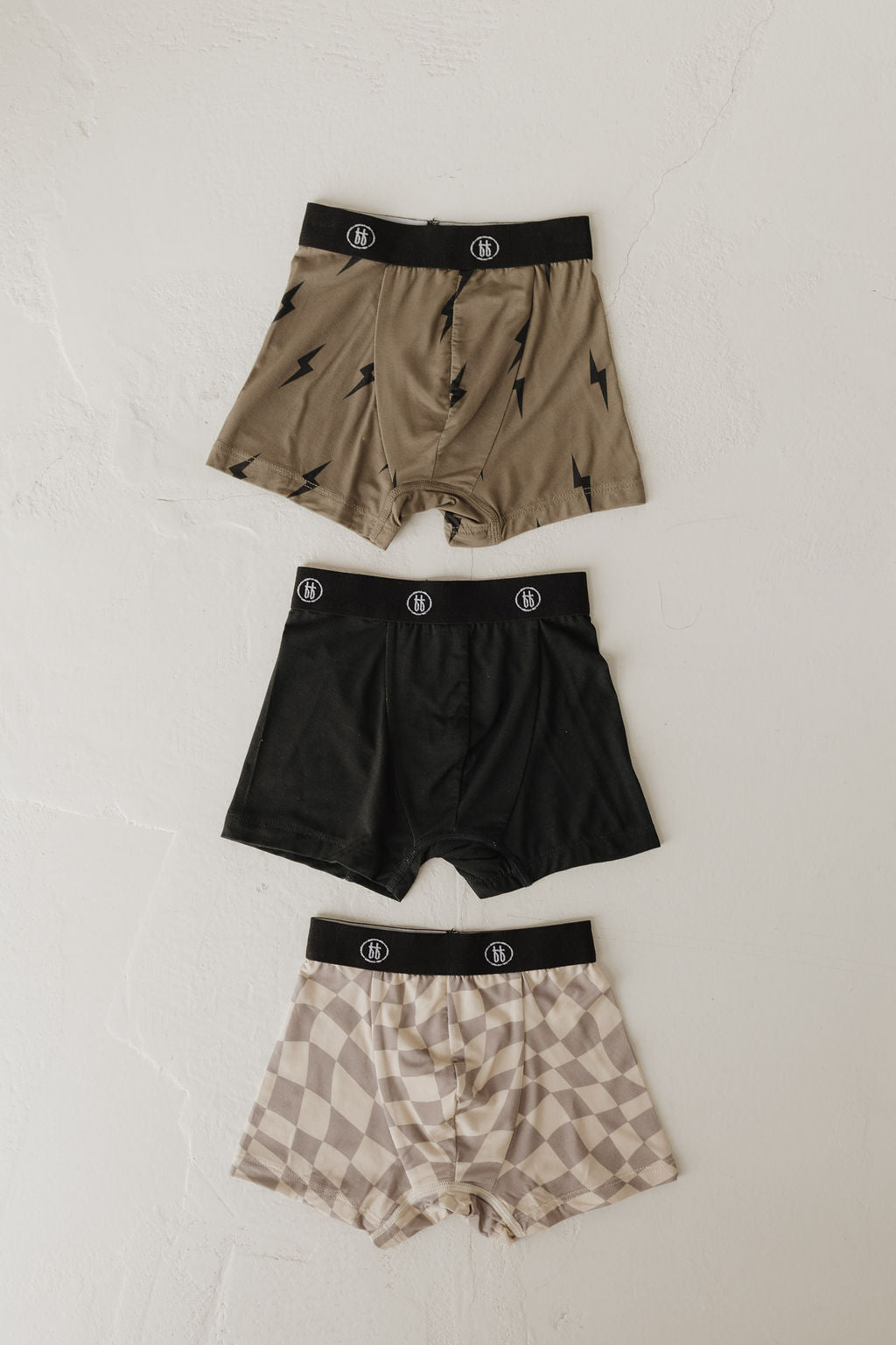 Three pairs of men's Bamboo Boxers from forever french baby are displayed. The top pair has an olive green with black lightning bolt design, the middle is solid black, and the bottom features a beige and white checkerboard pattern, all crafted from breathable bamboo fabric with black waistbands.