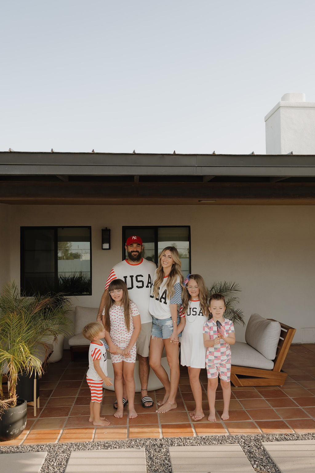 A family of six poses on a patio, wearing forever french baby’s breathable Bamboo T-Shirt Dress | Stars & Stripes and patriotic outfits. Two adults sport "USA" shirts, while four kids smile happily, some waving small American flags.