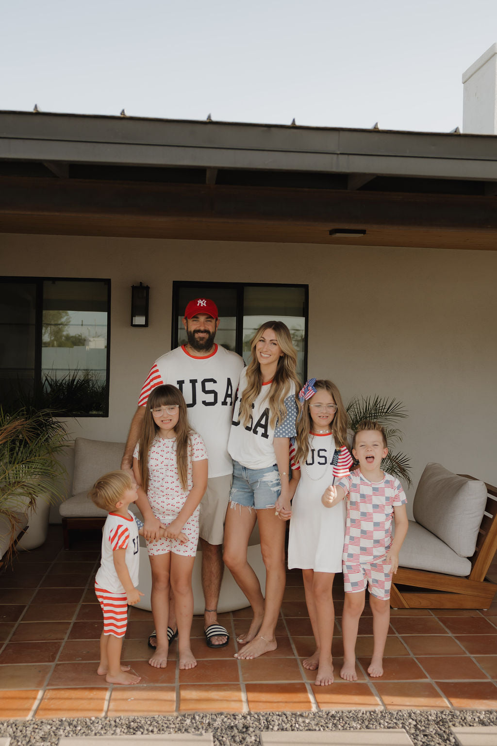 A family of five enjoys a sunny patio. The parents wear matching USA shirts, while the kids sport forever french baby Bamboo T-Shirt Dress | Stars & Stripes—breathable, hypo-allergenic pajamas perfect for festive summer fun.