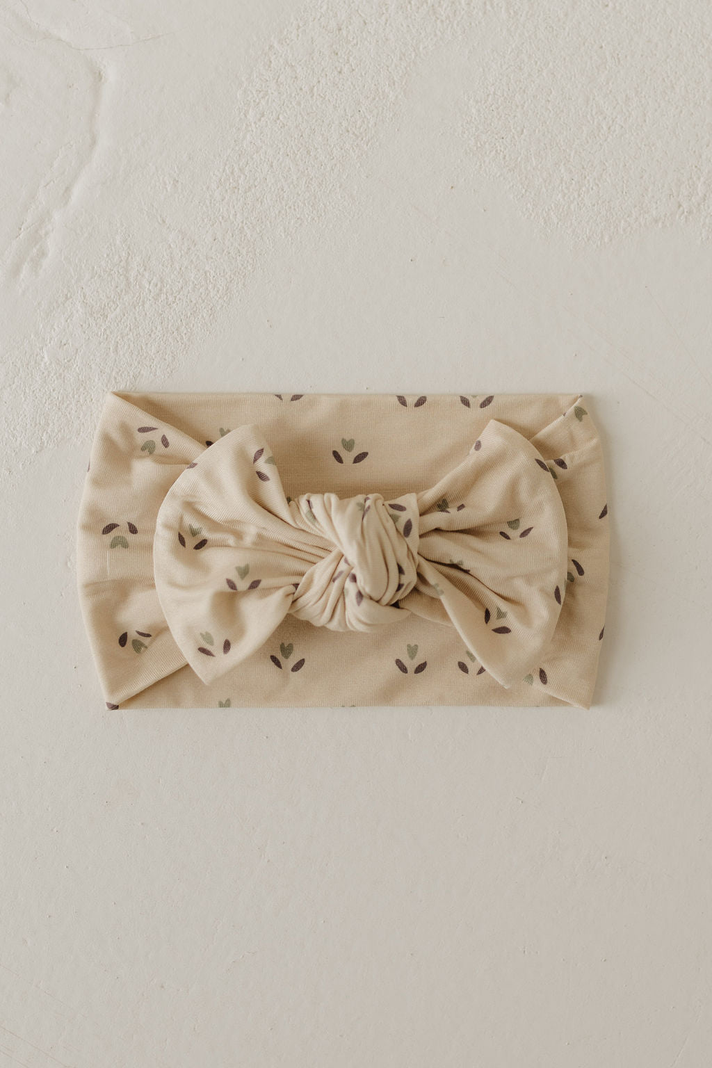 The Bamboo Head Wrap | Flora by forever french baby features a large central bow and a subtle brown leaf pattern, elegantly set against a textured, light-colored backdrop.
