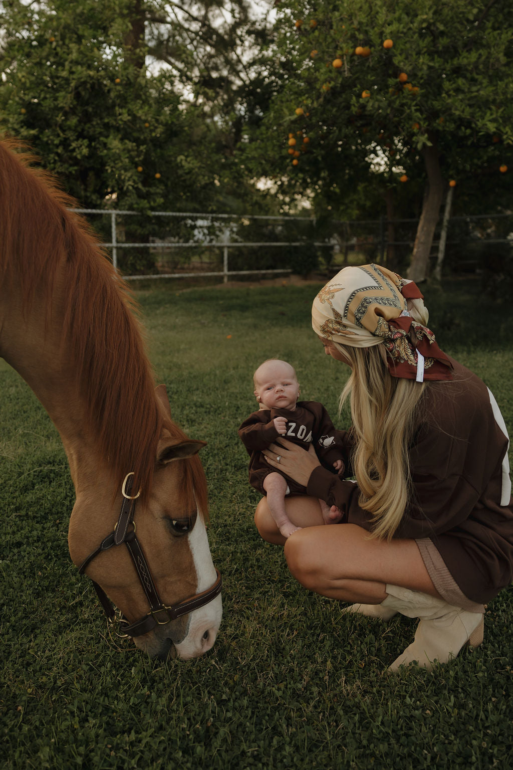 A woman in a headscarf kneels on the grass holding a baby wearing the forever french baby Sweatshirt Romper | ff Arizona, as a brown horse with a white stripe grazes nearby. Orange trees and a fence create a peaceful outdoor setting.