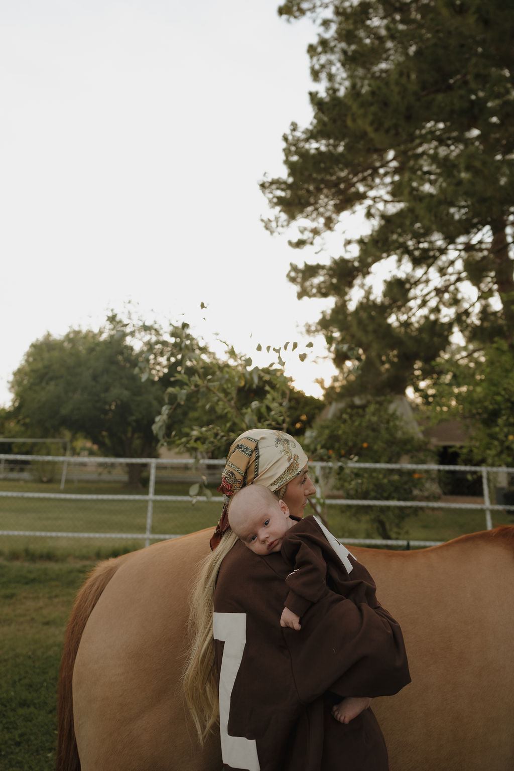 A woman in a bandana and brown Sweatshirt Romper | ff Arizona by forever french baby holds a baby, standing next to a light brown horse in a grassy yard with trees and a fence in the background.