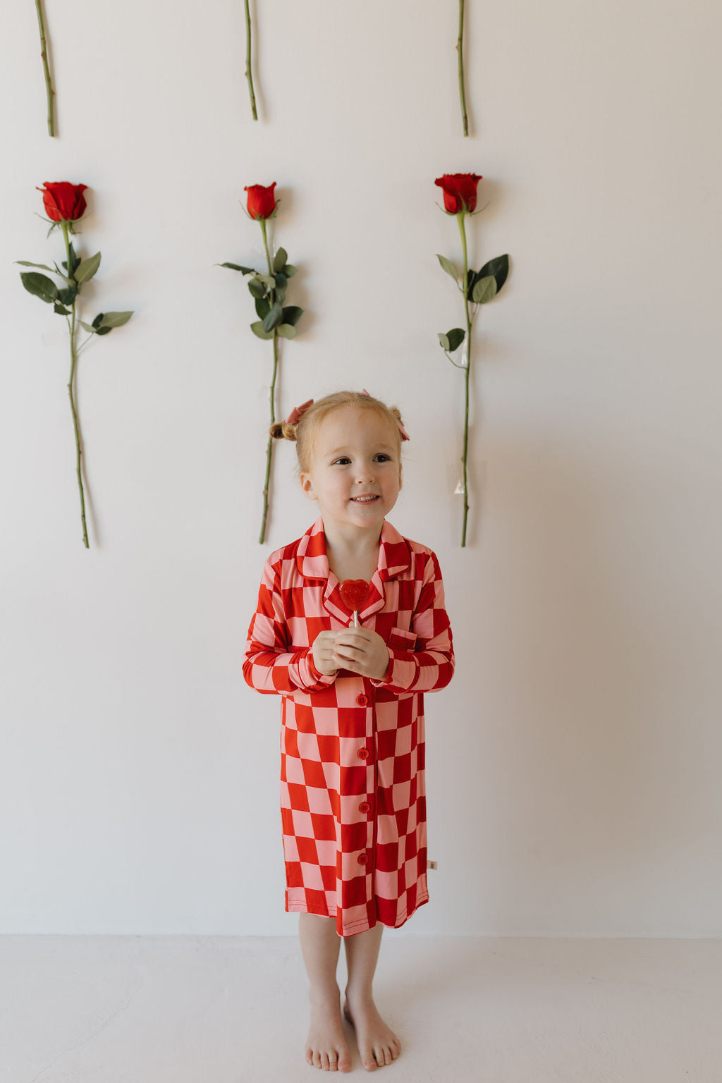 A young girl in a red and white checkered "Toddler Bamboo Sleeping Dress | XOXO" from forever french baby stands barefoot against a white wall, holding a red rose. Five red roses are vertically aligned on the wall behind her. She has braided hair and a cheerful expression.