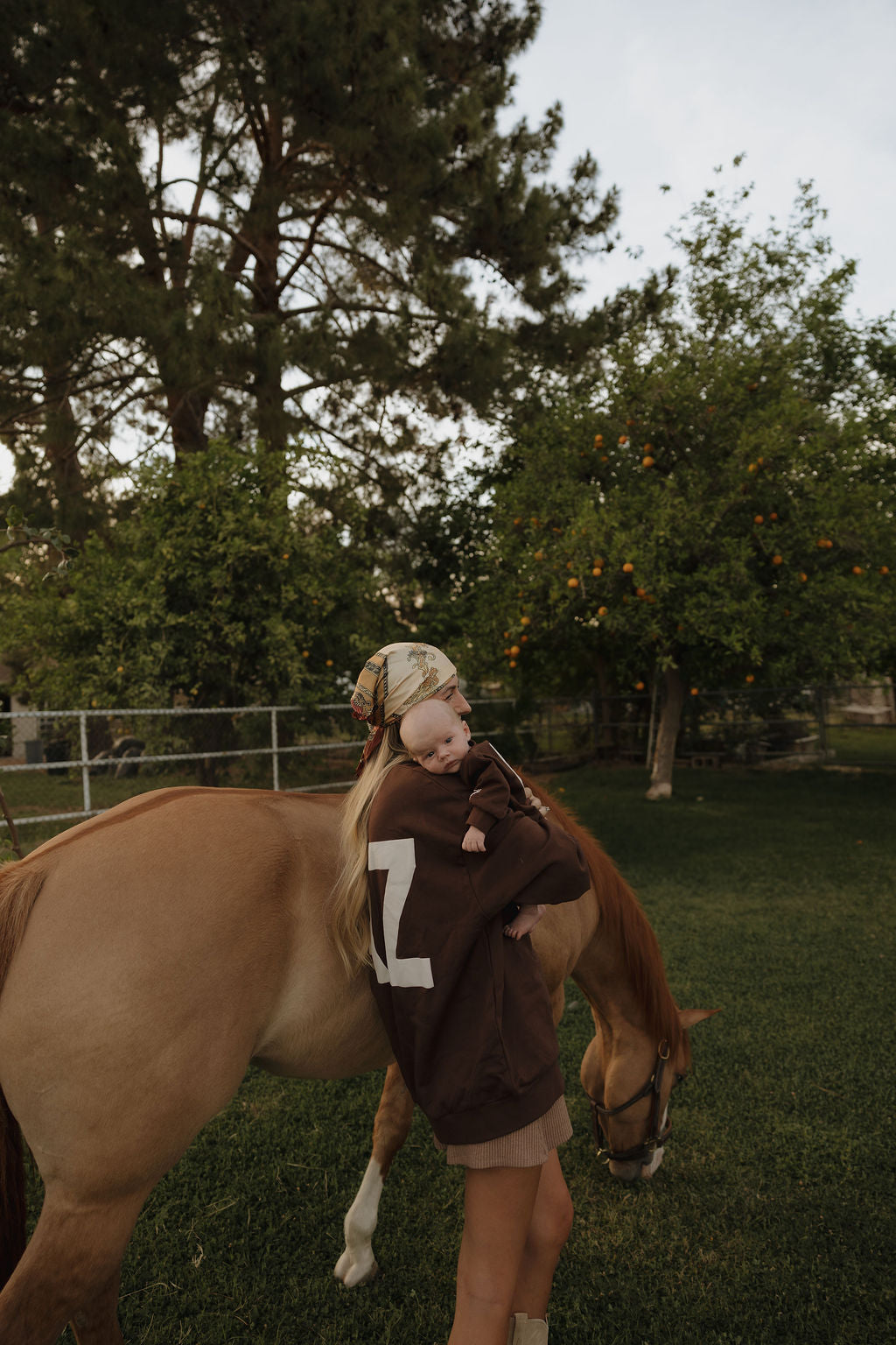 A woman in a relaxed fit ff Arizona Adult Sweatshirt by forever french baby and a headscarf holds a baby beside a light brown horse grazing in a green, tree-filled yard.