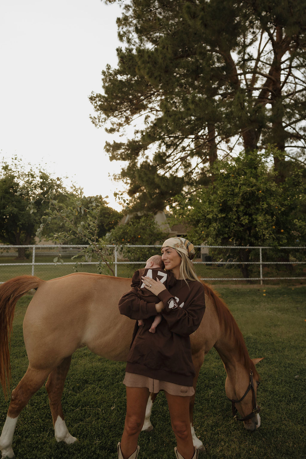 A woman in matching family outfits stands on grass holding a baby, with a light brown horse grazing nearby. The scene features trees and fencing, perfect for highlighting the forever french baby Sweatshirt Romper | ff Arizona.