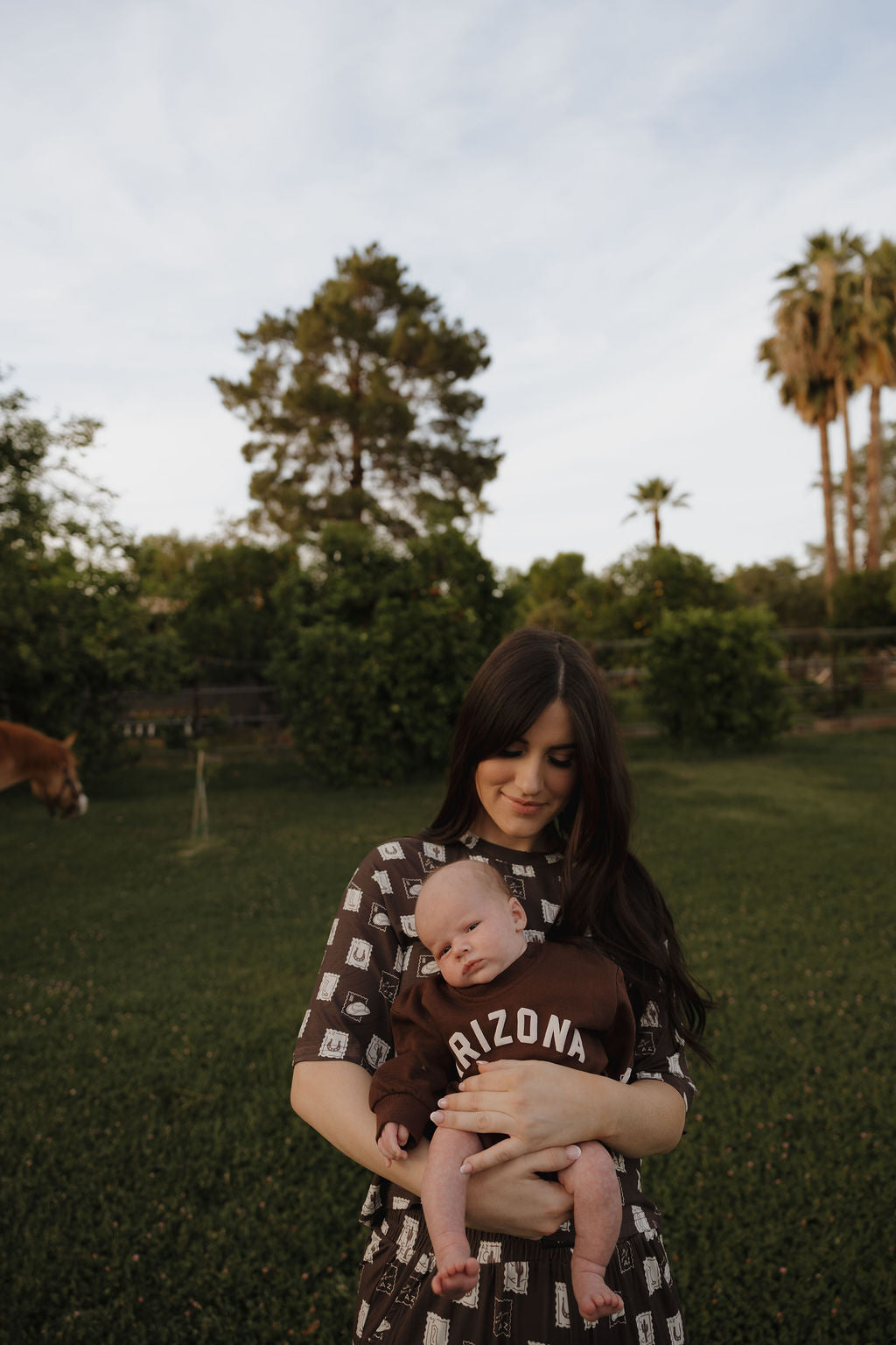 A woman in a patterned dress stands on grass holding a baby wearing the "Sweatshirt Romper | ff Arizona" by forever french baby. Outdoors with trees and palms, they’re ready for adorable matching family outfits on a sunny day.