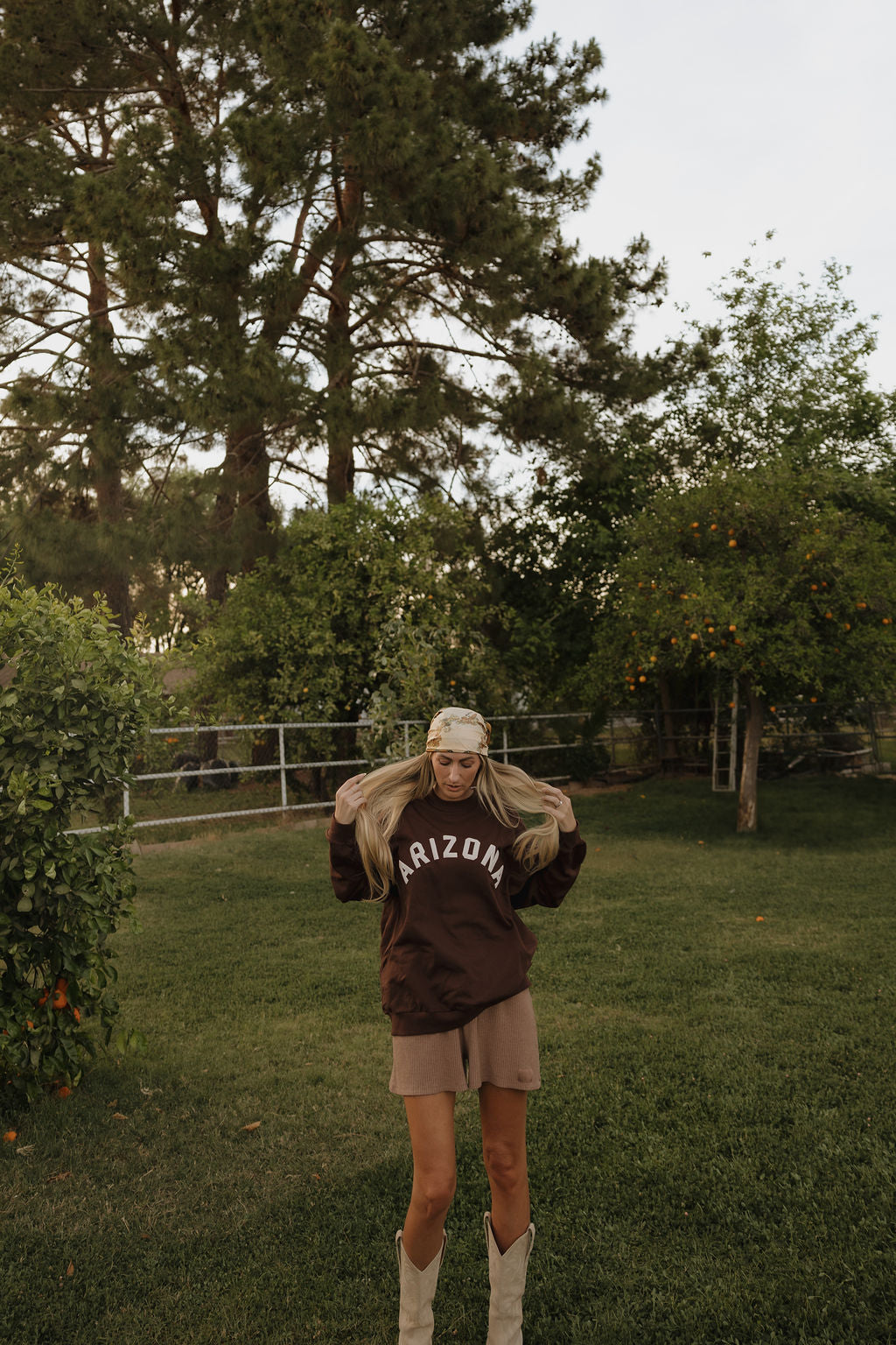 A woman stands on grass by trees, wearing a relaxed fit, 100% cotton Adult Sweatshirt | ff Arizona by forever french baby, paired with shorts, a bandana, and cowboy boots. She holds her long blonde hair with both hands.