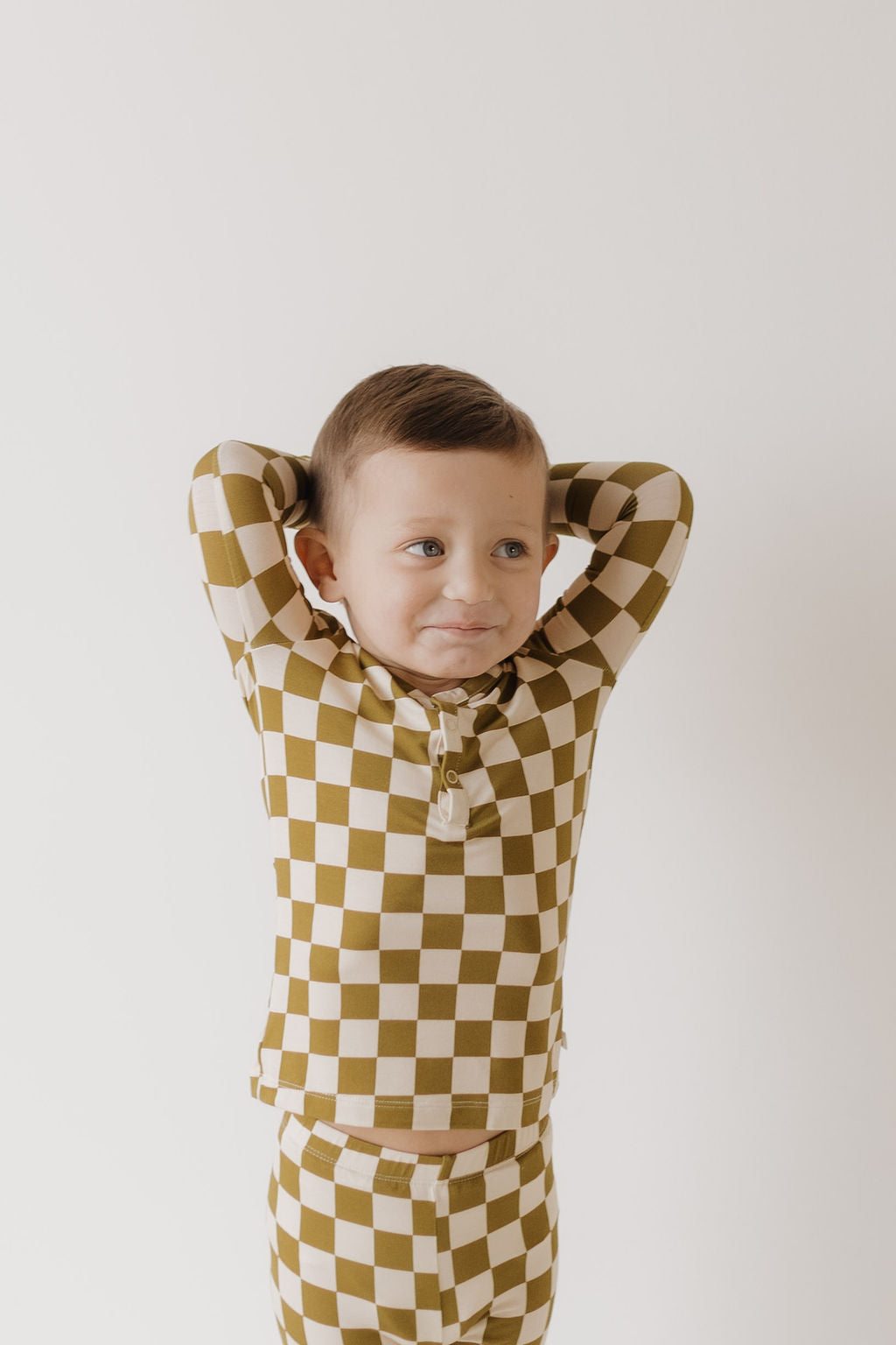 A child smiles with hands on their head while wearing breathable, hypoallergenic Olive Checkerboard Bamboo Two Piece Pajamas by forever french baby, featuring a long-sleeve top and pants against a neutral background.