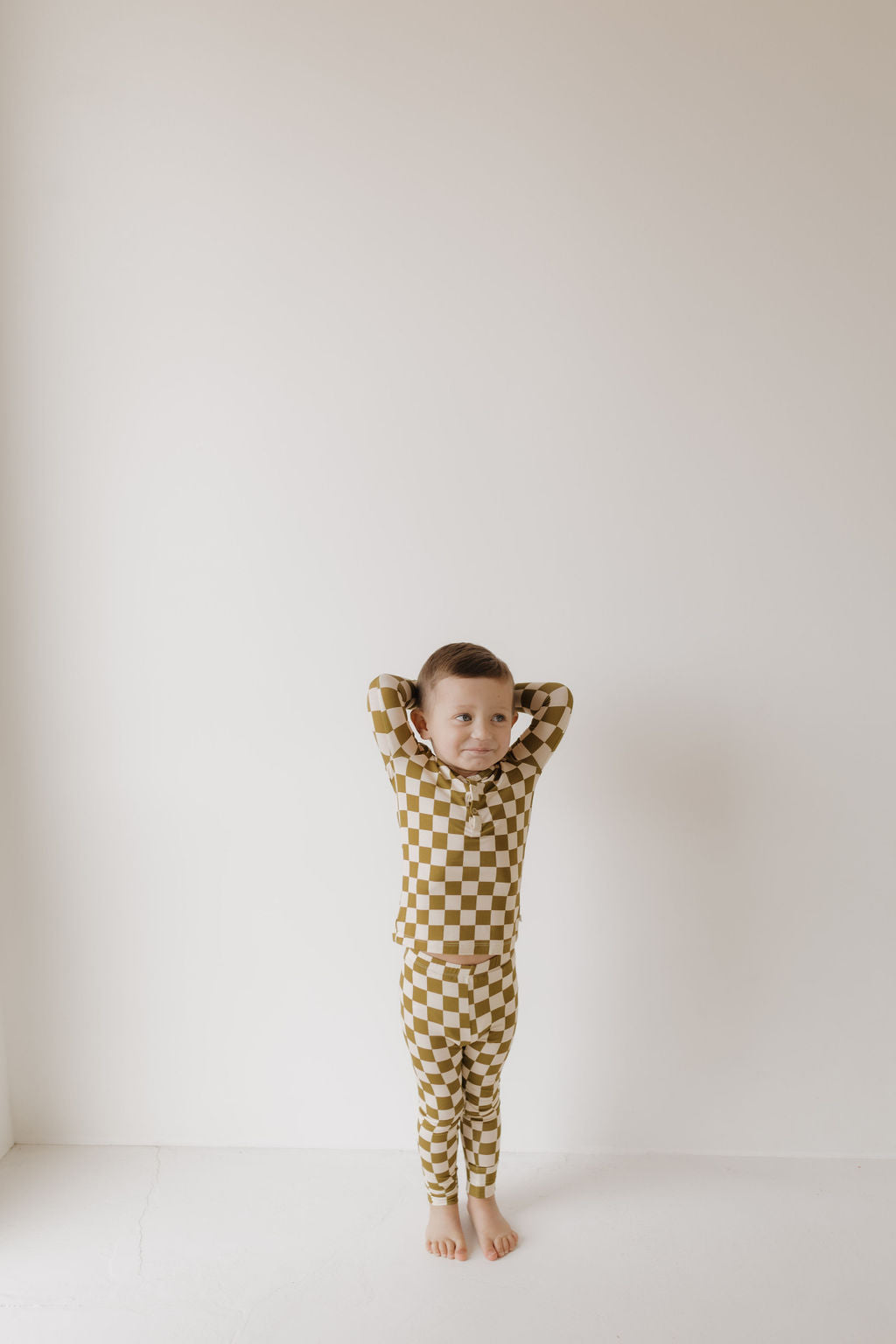 A young child stands barefoot against a white wall, smiling gently while wearing forever french baby's Bamboo Two Piece Pajamas in Olive Checkerboard. Hypoallergenic for comfort and style, they rest their hands behind their head.