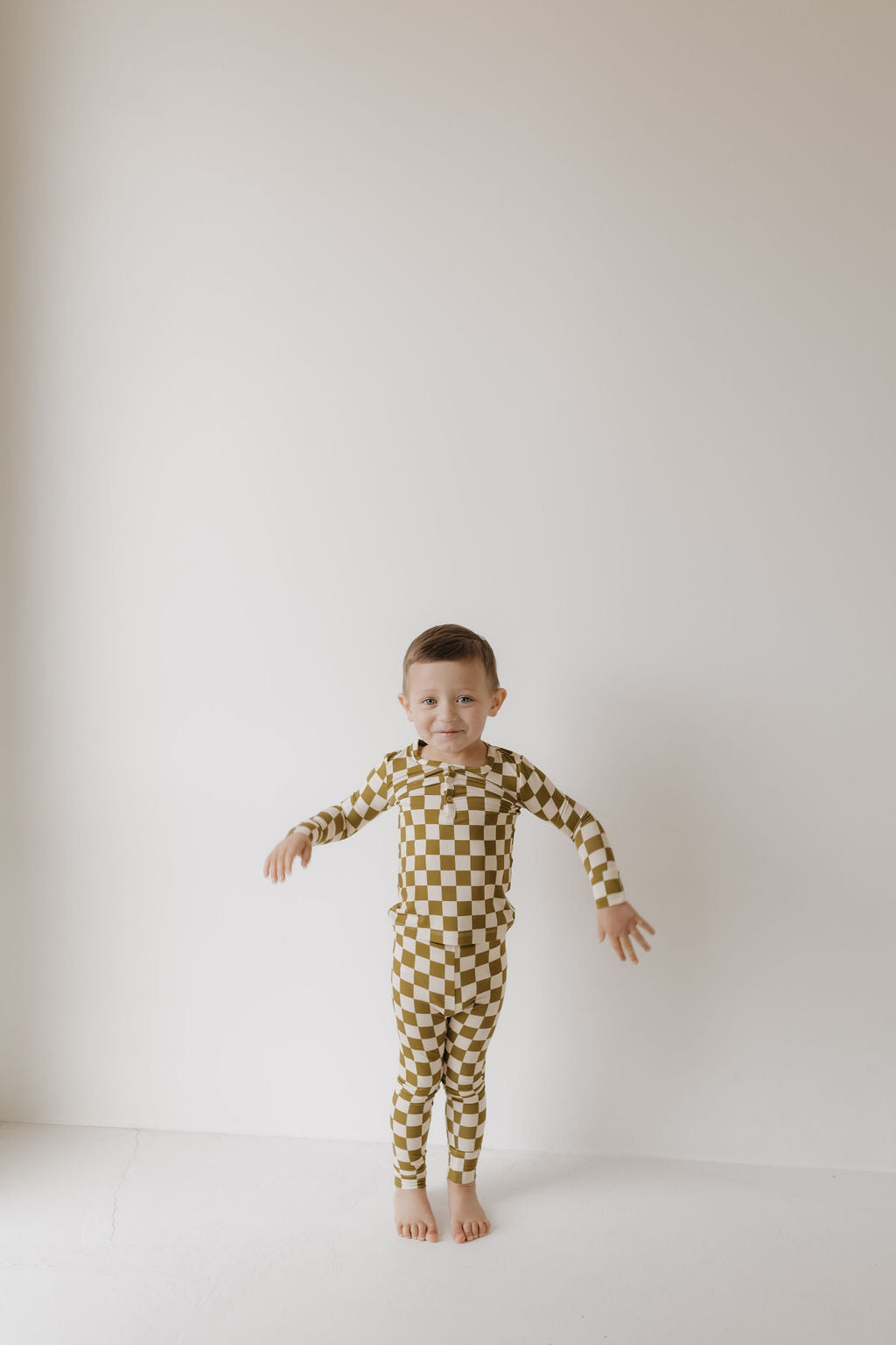 A young child is seen barefoot against a plain white backdrop, wearing forever french baby's Bamboo Two Piece Pajamas in Olive Checkerboard. The short-haired child looks straight ahead with slightly raised arms, exuding comfort and ease.