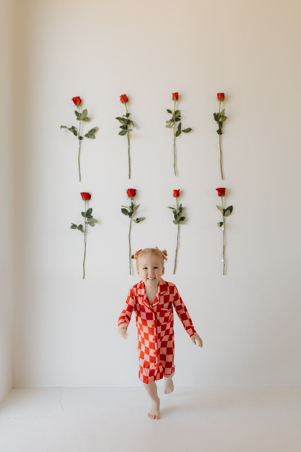 A little girl in a Toddler Bamboo Sleeping Dress from forever french baby smiles as she runs indoors. Behind her, nine red roses with green stems are arranged on a white wall in three rows. The room features minimalist and bright decor.