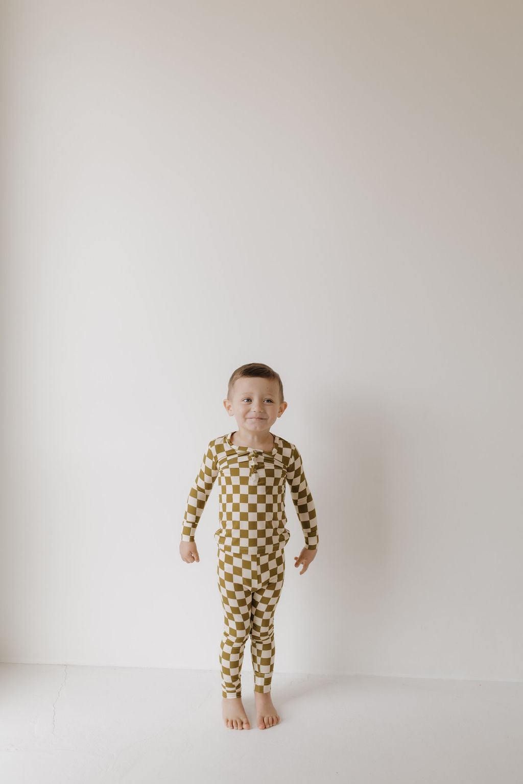 A child stands against a plain white wall in forever french baby's Bamboo Two Piece Pajamas in Olive Checkerboard. The hypo-allergenic outfit ensures comfort, while the barefoot child with short brown hair smiles subtly in the minimalistic, well-lit setting.