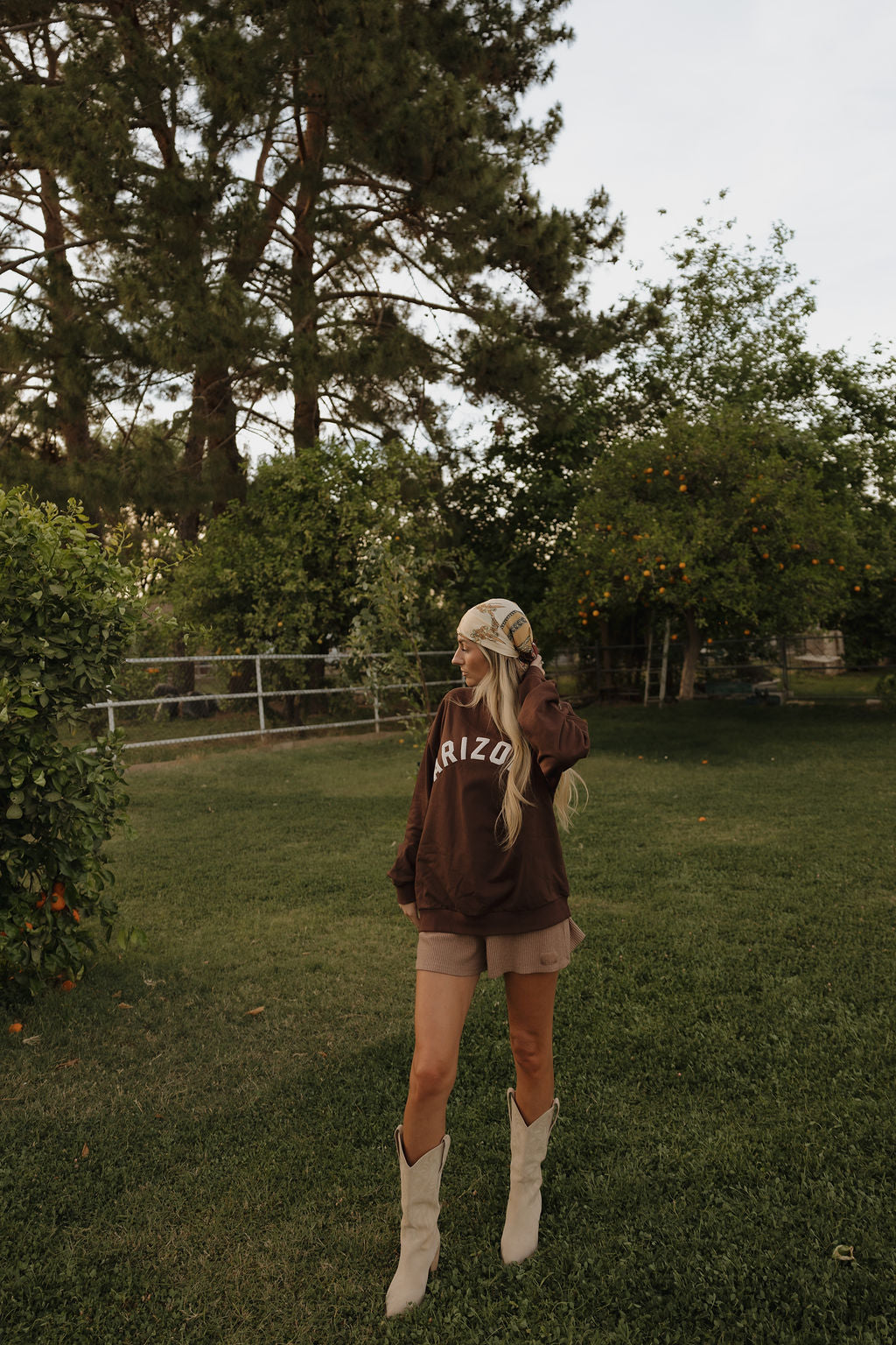 A woman in a relaxed fit, 100% cotton Adult Sweatshirt | ff Arizona by forever french baby, tan shorts, a patterned headscarf, and white cowboy boots stands on grass in a green yard with trees and orange bushes behind her.
