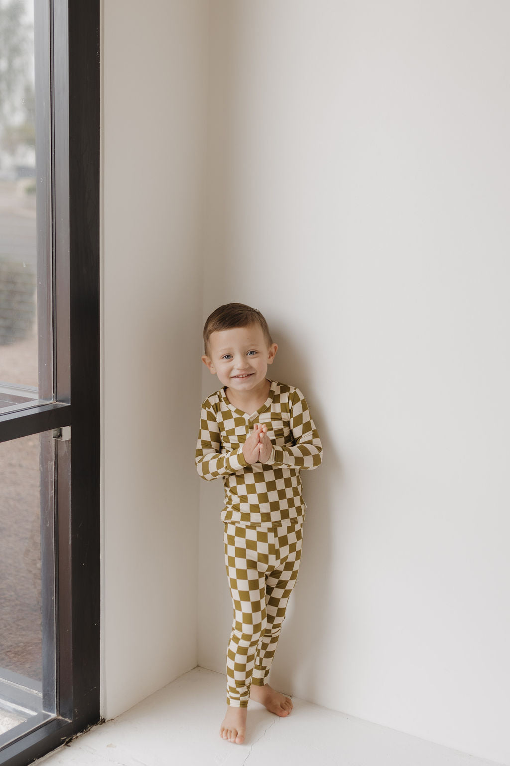 A young child stands by a window, barefoot and smiling in natural light, wearing the Olive Checkerboard Bamboo Two Piece Pajamas by forever french baby.