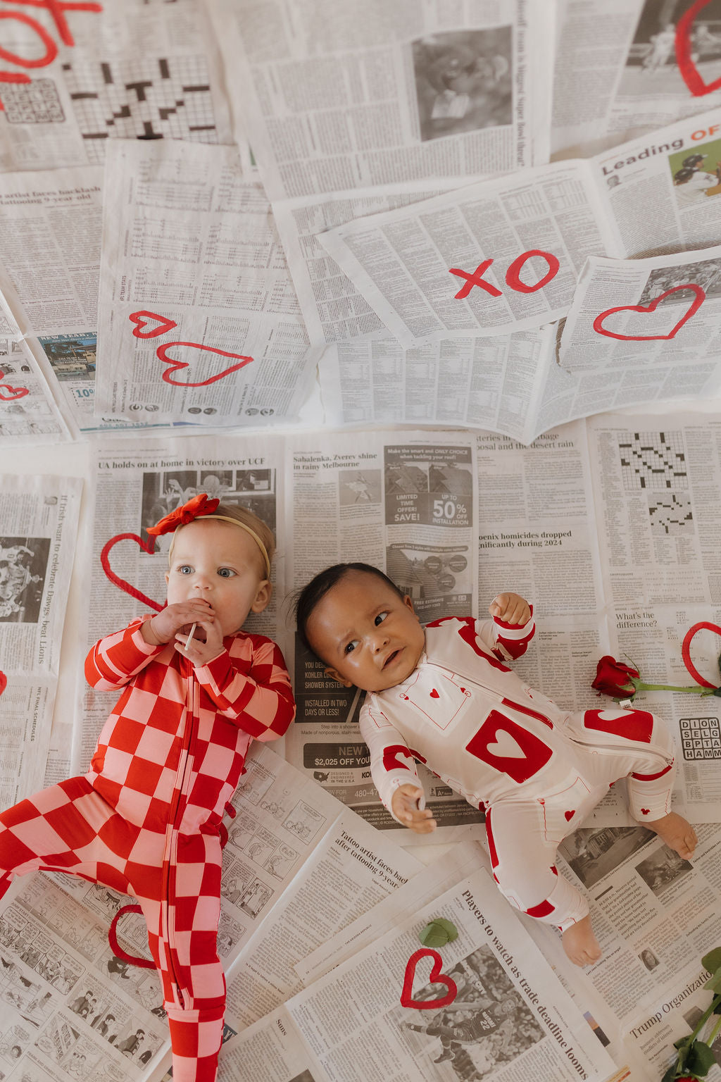 Two babies lie on a newspaper-covered floor wrapped in Forever French Baby's Bamboo Zip Pajamas | Love Day. One wears a red checkered outfit, and the other sports white with red hearts. "XO" and hearts adorn the newspapers, creating a cozy, playful atmosphere with hypo-allergenic comfort.