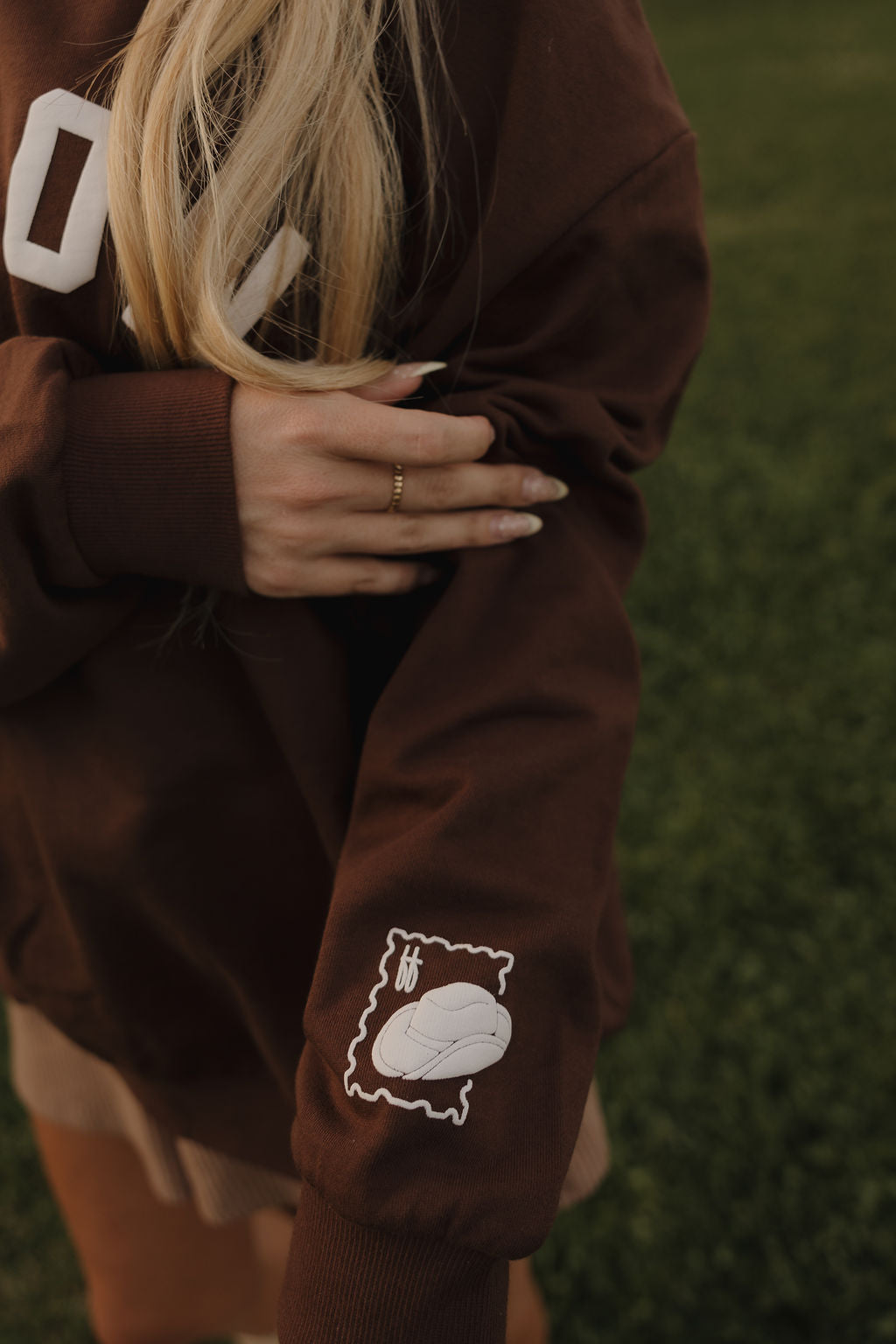 A person with long blonde hair stands on green grass wearing a relaxed fit brown "Adult Sweatshirt | ff Arizona" by forever french baby, featuring a rugby ball patch in a stamp-like border on the sleeve.
