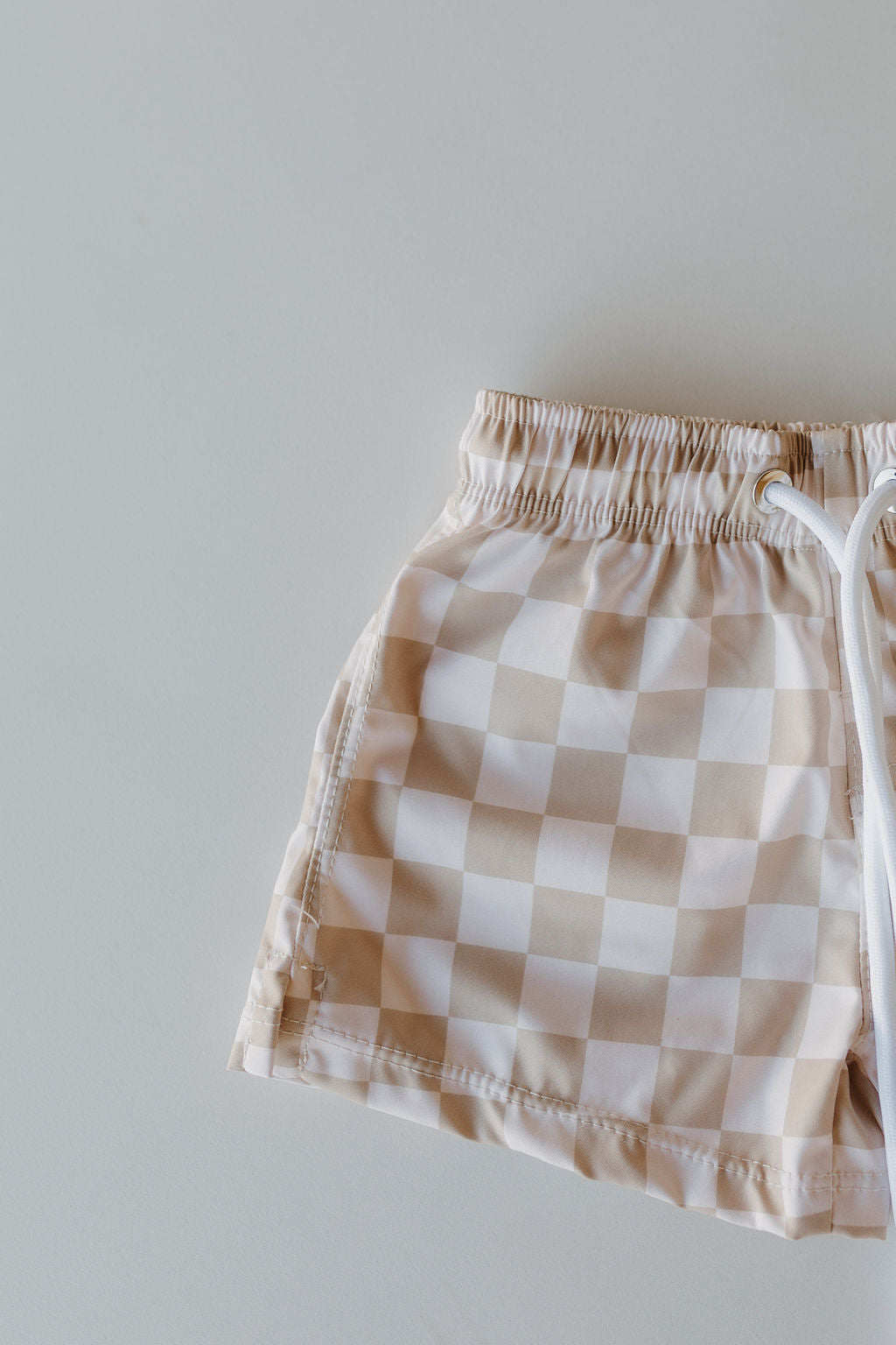 The Child Boardshort from the Forever French Baby Coastline collection, featuring a beige and white checkered design with an elastic waistband and white drawstrings, elegantly rests on a light gray surface.