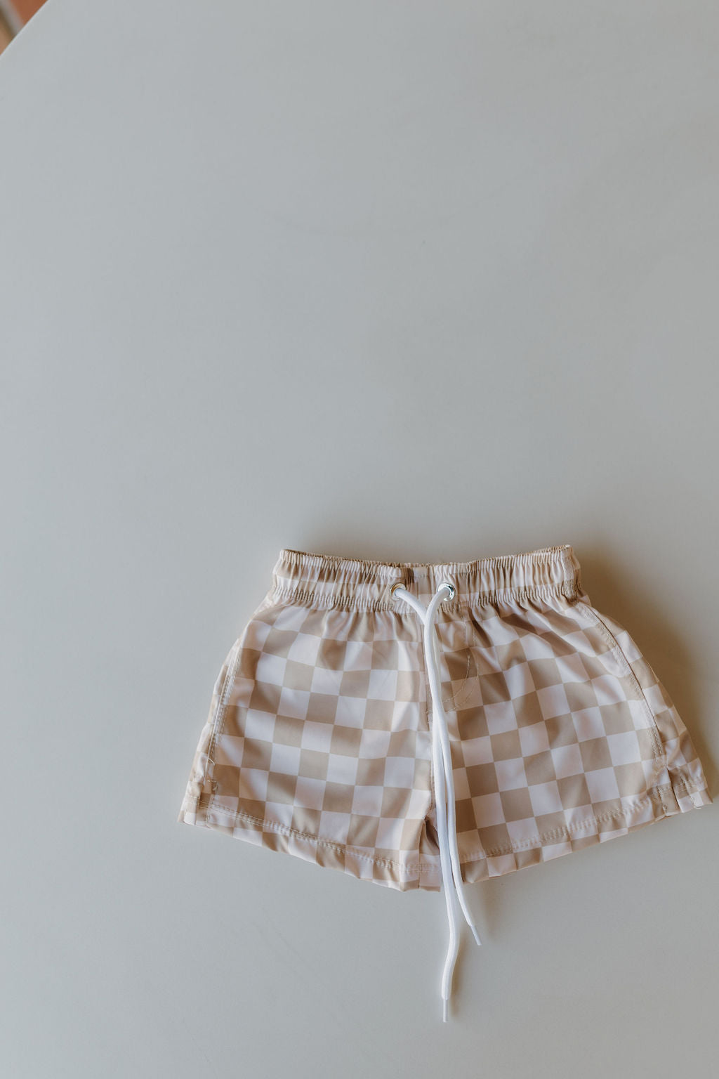 The Child Boardshort from the Coastline collection by forever french baby features a beige and white checkered pattern, a white drawstring, and an elastic waistband, shown on a light grey surface.