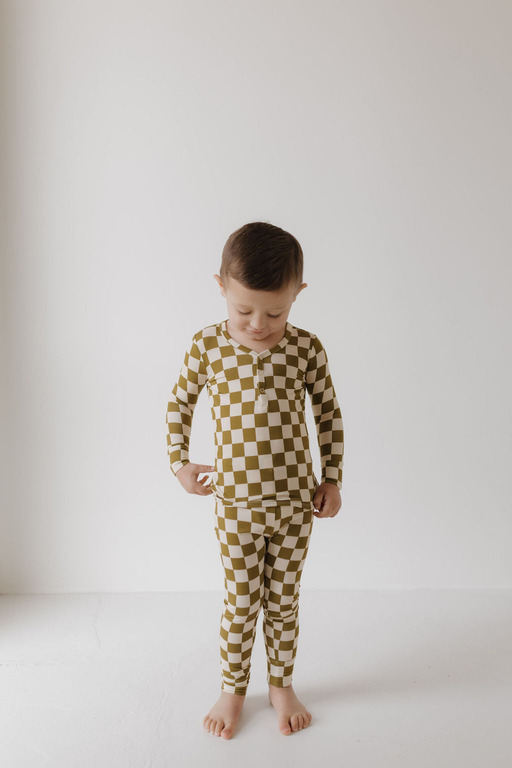 A young child stands barefoot against a plain backdrop, wearing Forever French Baby's Bamboo Two Piece Pajamas in Olive Checkerboard. The breathable fabric ensures their comfort as they look down with their hands by their sides in the softly lit room.