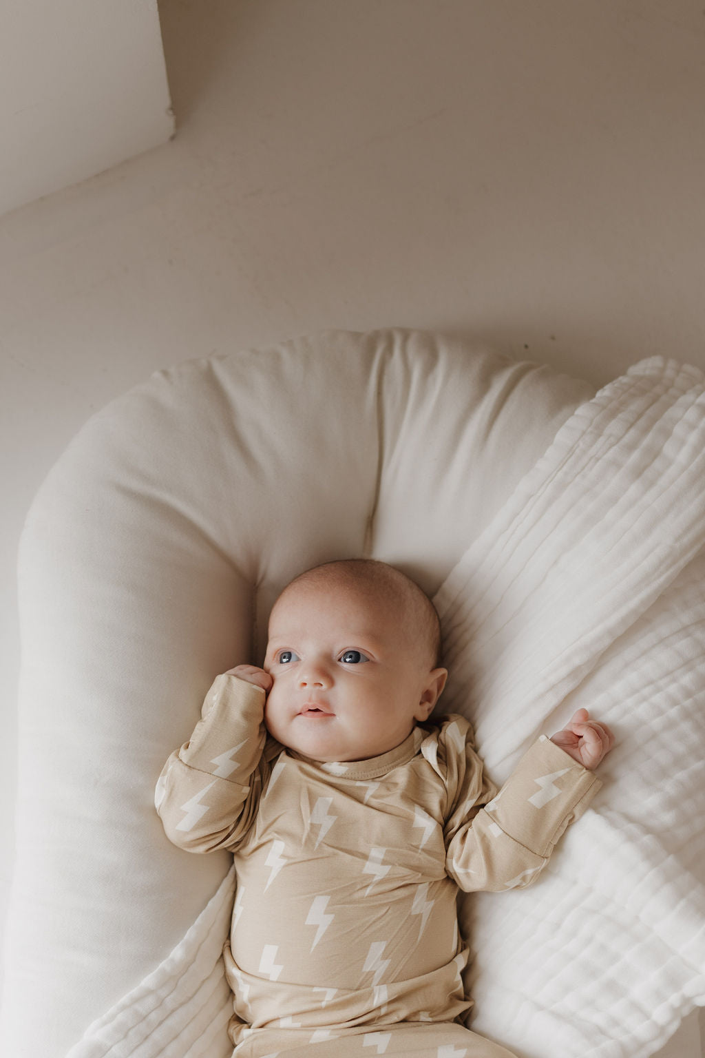 A baby rests on a soft white cushion in a "Bamboo Knotted Gown | Tan Lightning Bolt" from forever french baby, looking up while partially covered by a hypoallergenic blanket—truly essential for newborns.