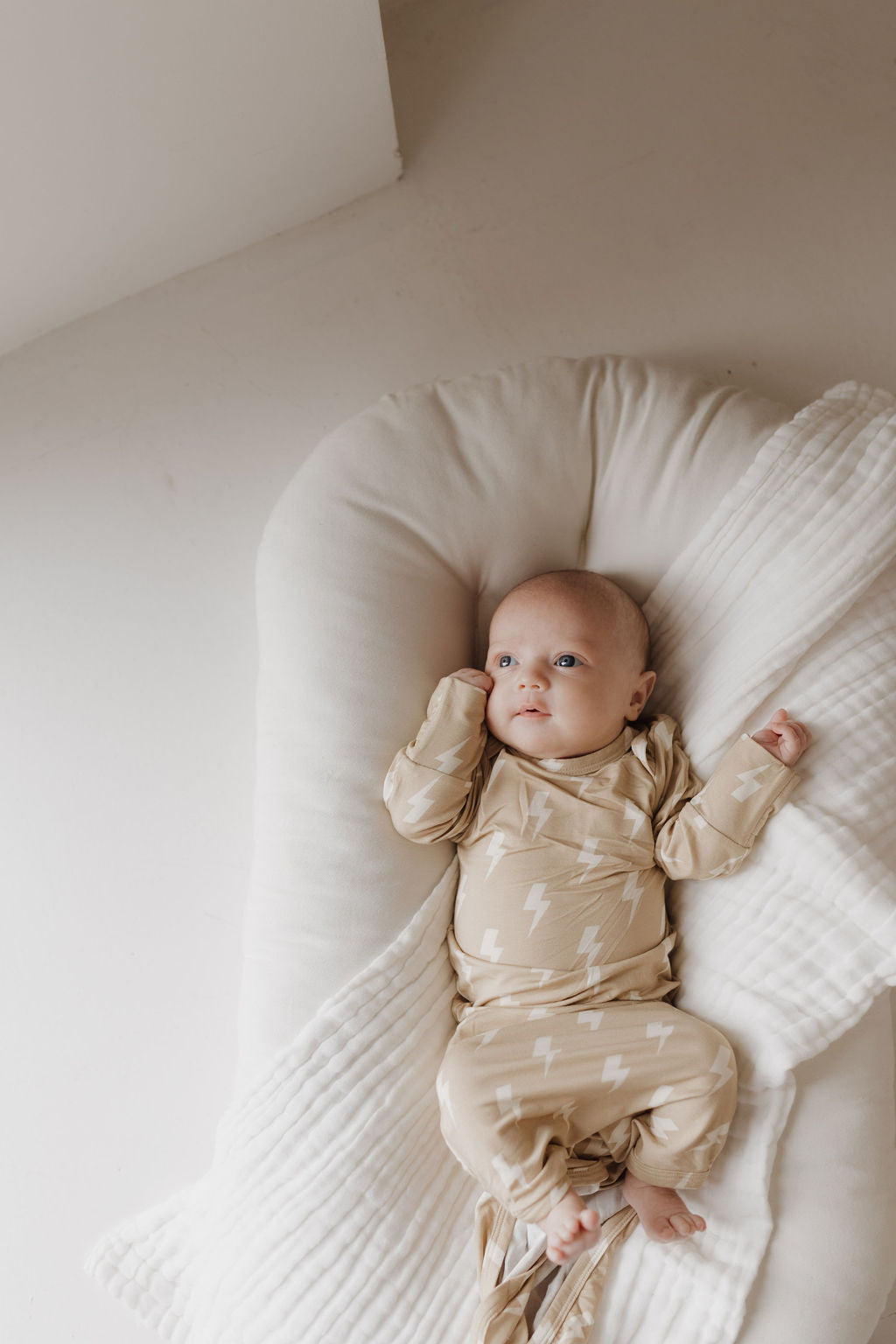 A baby in a Bamboo Knotted Gown by forever french baby, featuring a tan lightning bolt pattern, lies on a soft white cushion. The hypoallergenic bamboo outfit adds to the serene ambiance as the baby gazes upward with one arm bent near their head under gentle lighting.