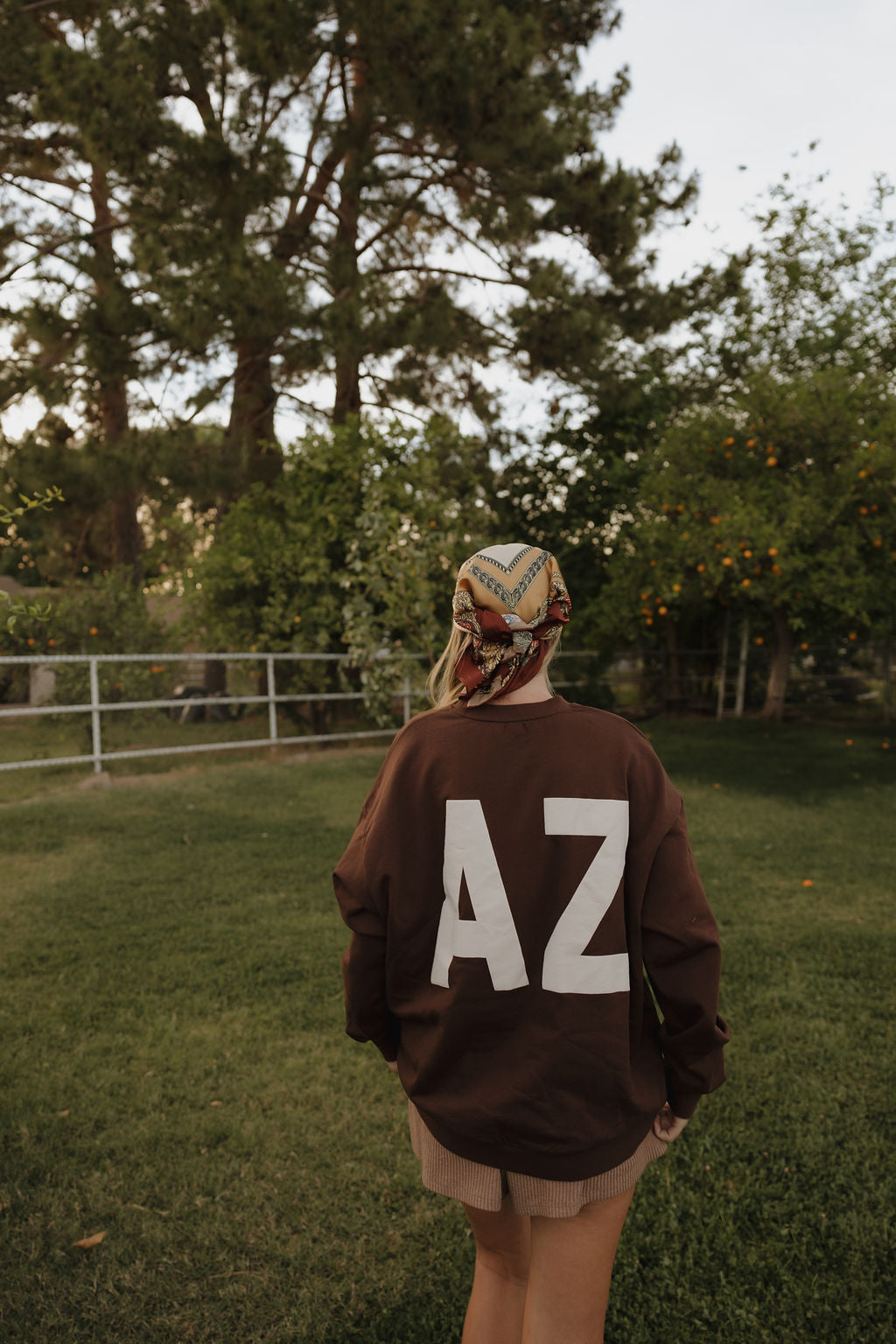Wearing a relaxed fit, 100% cotton Adult Sweatshirt | ff Arizona by forever french baby with bold white "AZ" lettering, a person in a patterned headscarf stands on grass in a tree-lined yard, facing away from the camera.