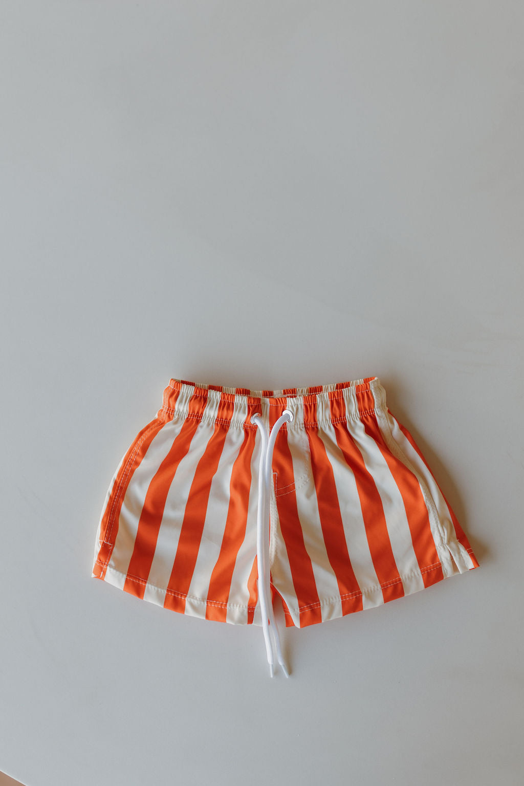 From the Poolside Collection by forever french baby, these Child Boardshorts feature orange and white vertical stripes, a white drawstring, and an elastic waistband. Perfect for summer or beach outings, they are displayed on a light gray surface.