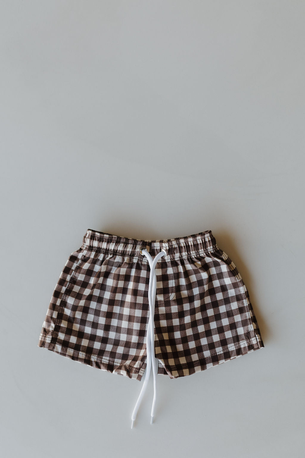 From the Chocolate Gingham collection by forever french baby, the Child Boardshort in brown and white checks features an elastic waistband and a white drawstring, styled flat on a neutral background.