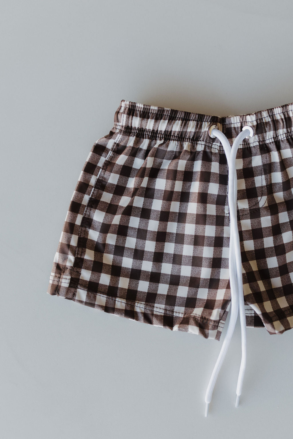 The Child Boardshort from forever french baby's Chocolate Gingham collection features a brown and white checkered pattern, an elastic waistband, and a white drawstring, shown flat on a light gray surface.