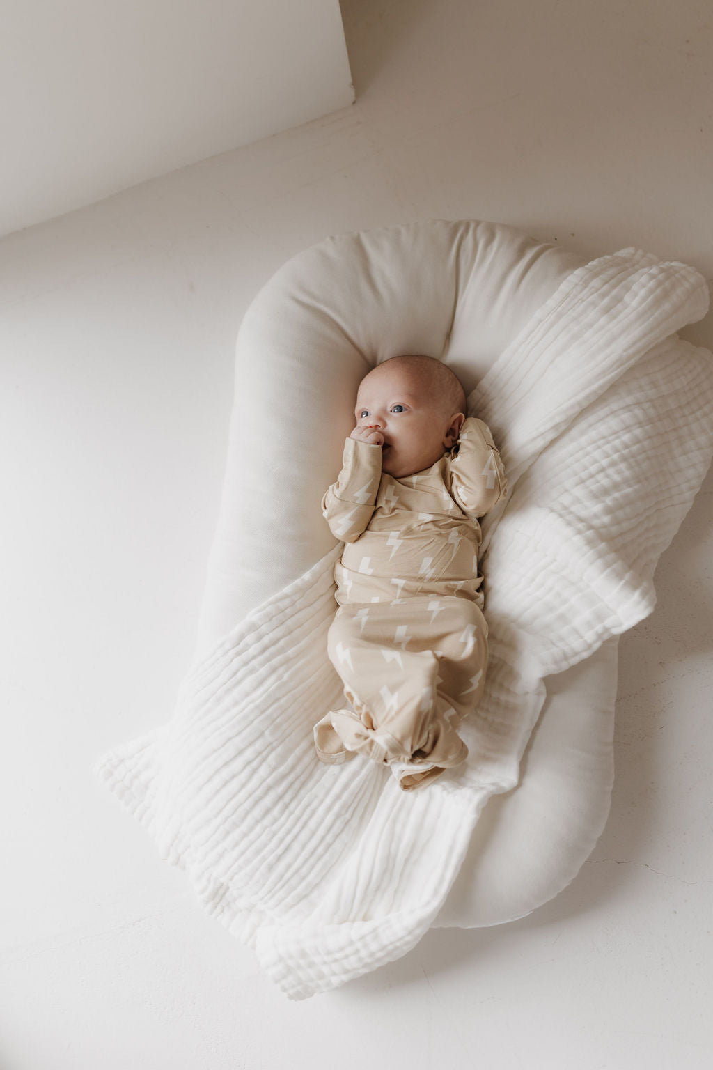 Wearing a "Bamboo Knotted Gown | Tan Lightning Bolt" by forever french baby, a baby lies on a cushioned white surface, sucking their thumb and partly covered by a textured white blanket in a bright, minimalist setting.