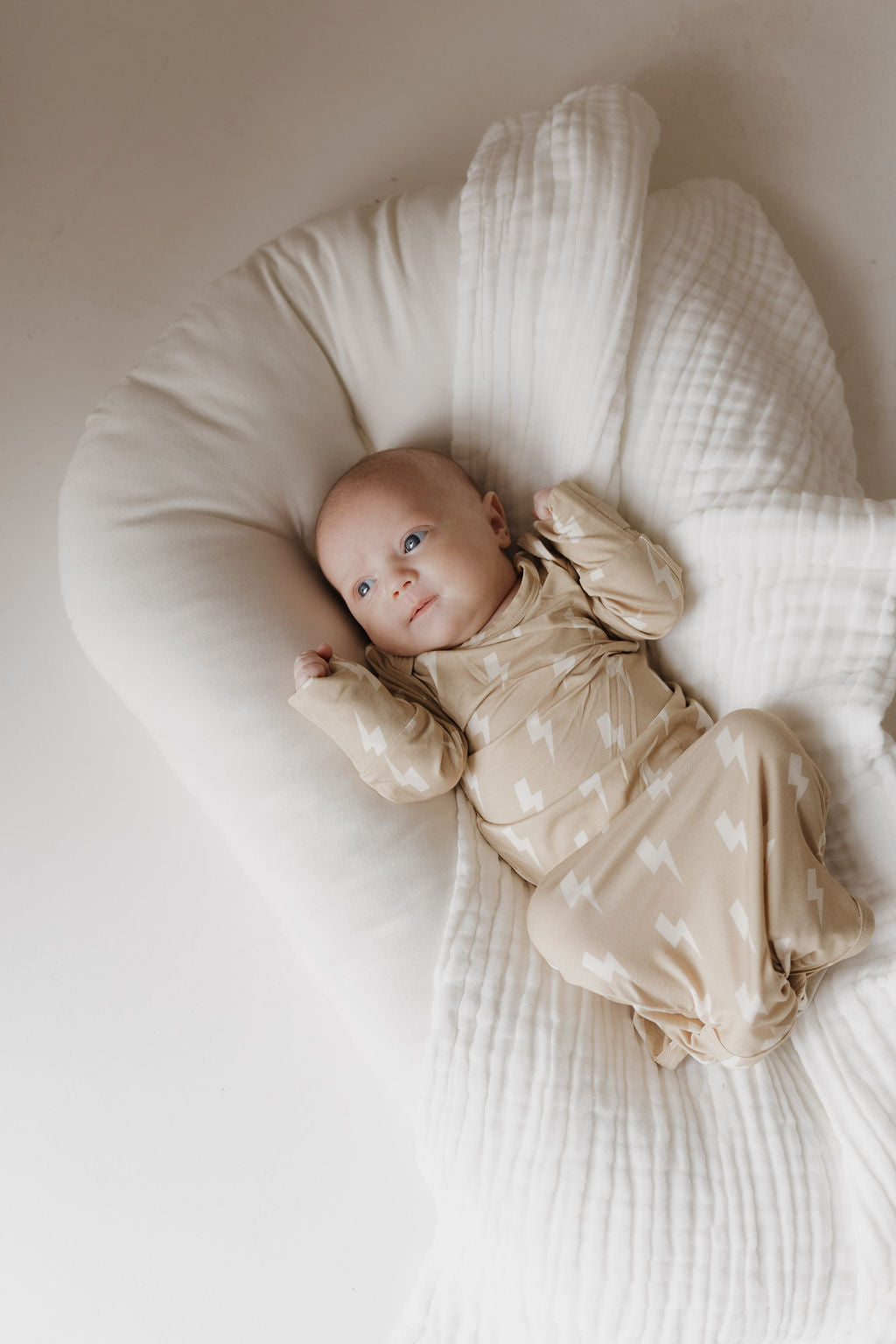 A baby, wrapped in a hypo-allergenic swaddle with small zigzags and dressed in a "Bamboo Knotted Gown | Tan Lightning Bolt" by Forever French Baby, lies on soft white fabric, gazing calmly against the gentle neutral background.