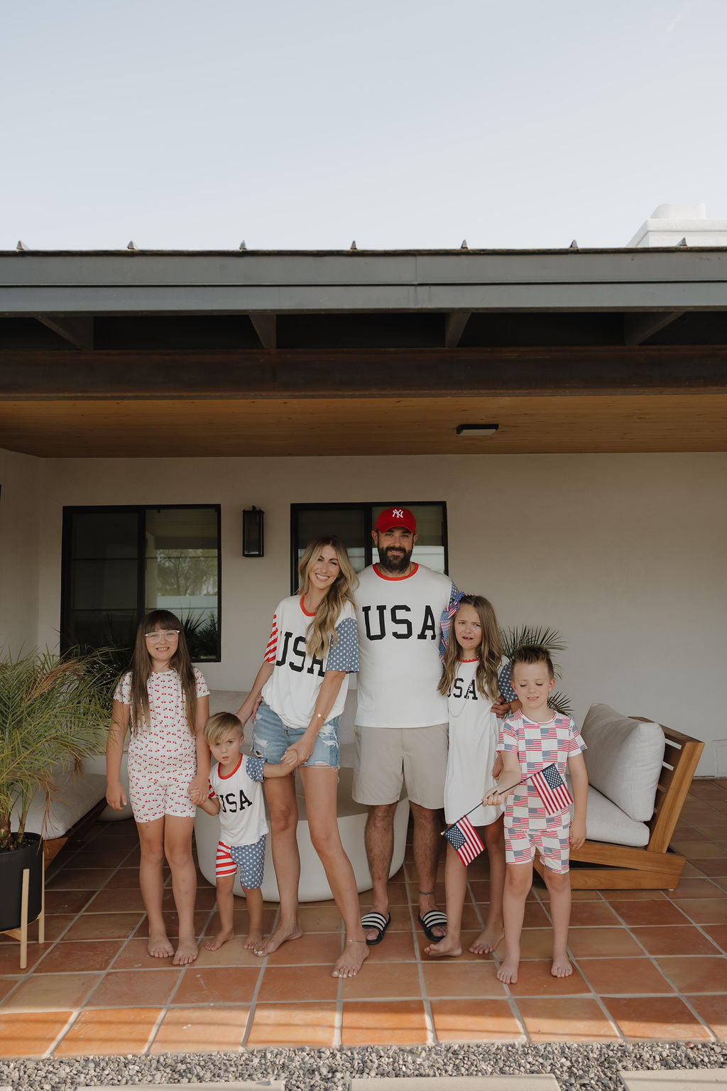 A family of six poses on their patio in matching forever french baby Bamboo T-Shirt Dress | Stars & Stripes outfits. Two kids smile and wave small American flags, showing off the breathable, USA-themed bamboo clothing.