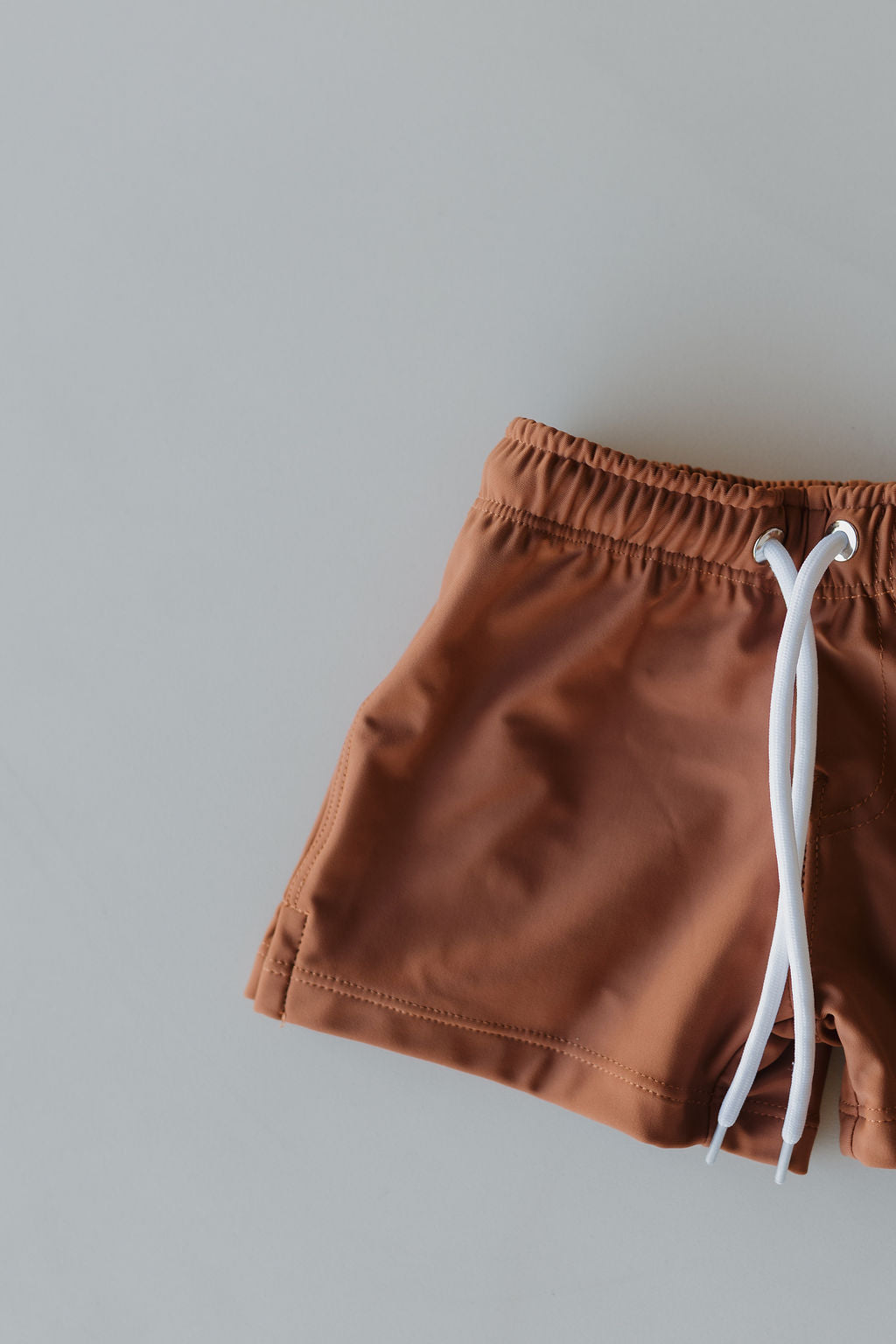 Child Boardshort | Terra by forever french baby, brown with a white drawstring, displayed on a light gray background. Only part of the shorts is shown, featuring an elastic waistband for a customizable fit and simple design.