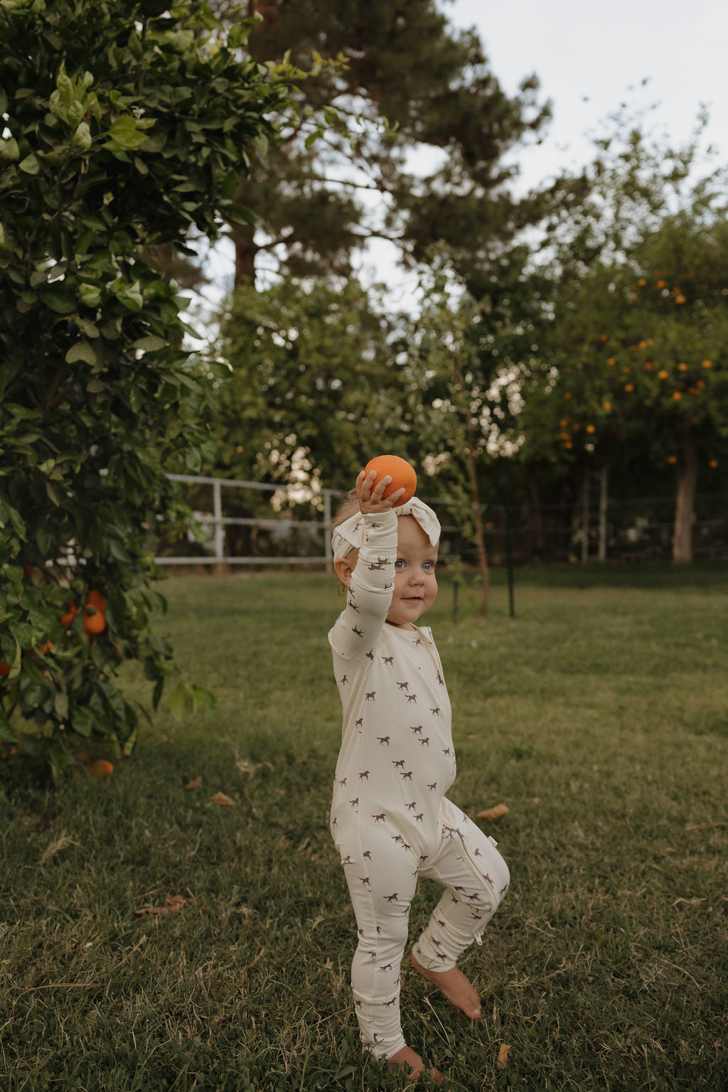A young child in forever french baby Bamboo Zip Pajamas | Wild Horses stands barefoot on grass, wearing a headband and holding an orange above their head in a garden with green trees and oranges in the background.