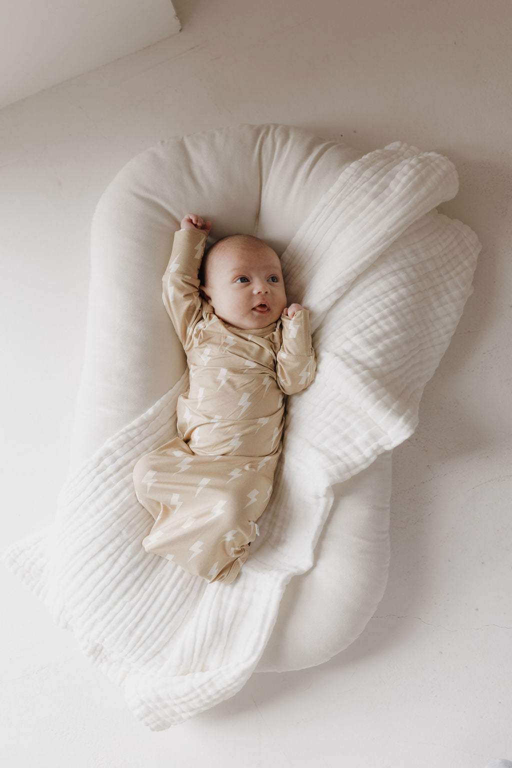 A baby rests on a cushioned white base, swaddled in the "Bamboo Knotted Gown | Tan Lightning Bolt" from forever french baby. Made of hypo-allergenic bamboo with a lightning bolt pattern, the newborn's raised arm and a partially covering white blanket create a cozy setting.