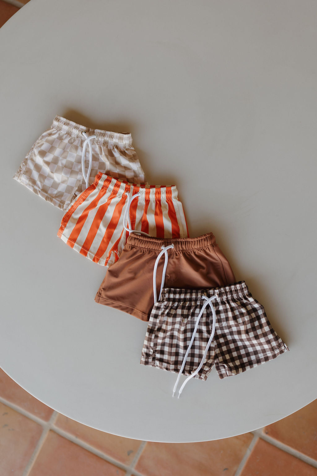 Four pairs of forever french baby Child Boardshort | Terra are arranged on a round table, showcasing designs from top to bottom: cream checkered, orange and white stripes, solid brown, and brown checkered. Each pair offers a customizable fit with a white drawstring.