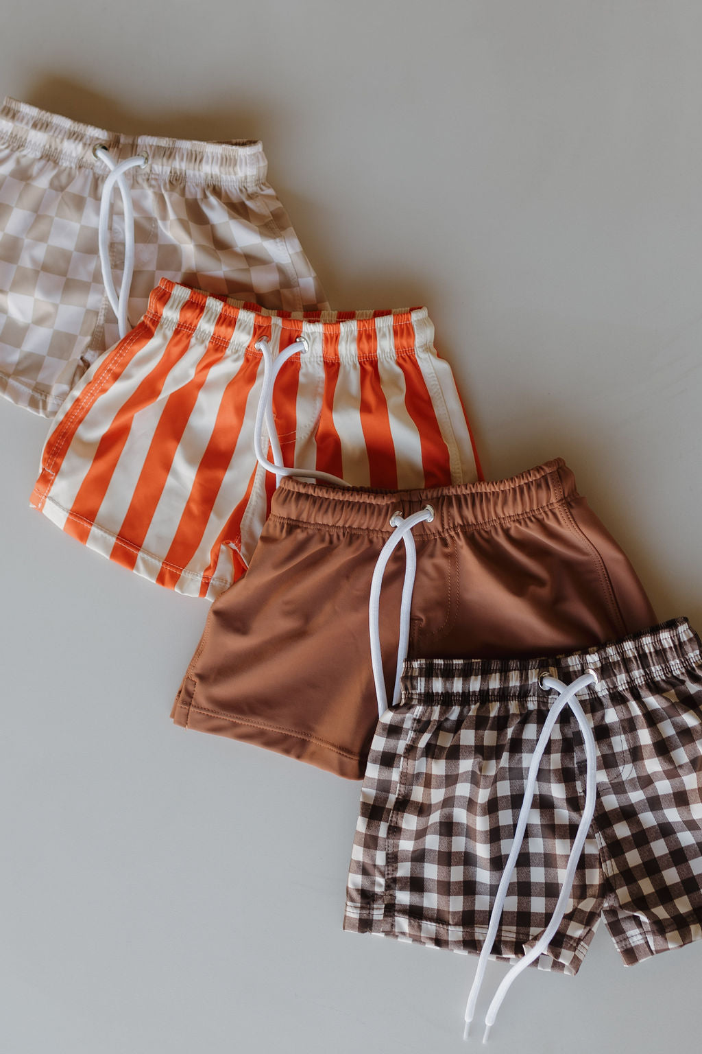 Four pairs of "Child Boardshort | Poolside" by Forever French Baby are displayed flat: beige checkered, orange and white striped, solid brown, and brown checkered. Ideal for kids' sunny pool days!.