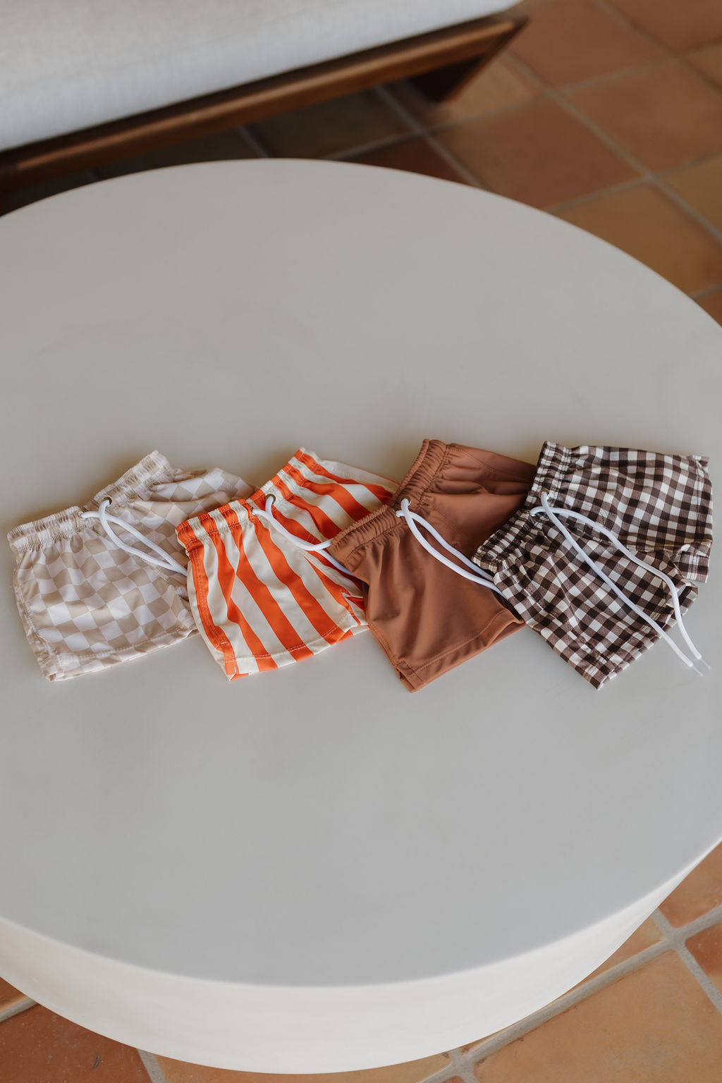 Four Child Boardshorts from forever french baby's Poolside Collection in beige checkered, orange striped, solid brown, and brown checkered designs with white drawstrings are stylishly arranged on a round table against a tiled floor backdrop.