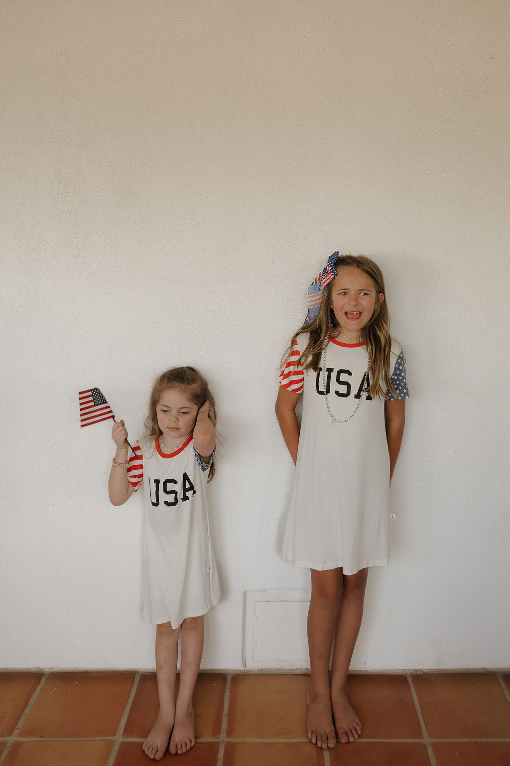 Two young girls stand barefoot against a plain wall, both wearing forever french baby Bamboo T-Shirt Dress | Stars & Stripes with "USA" printed on the front. The older girl smiles and holds a flag, while the younger looks down, holding a small American flag.
