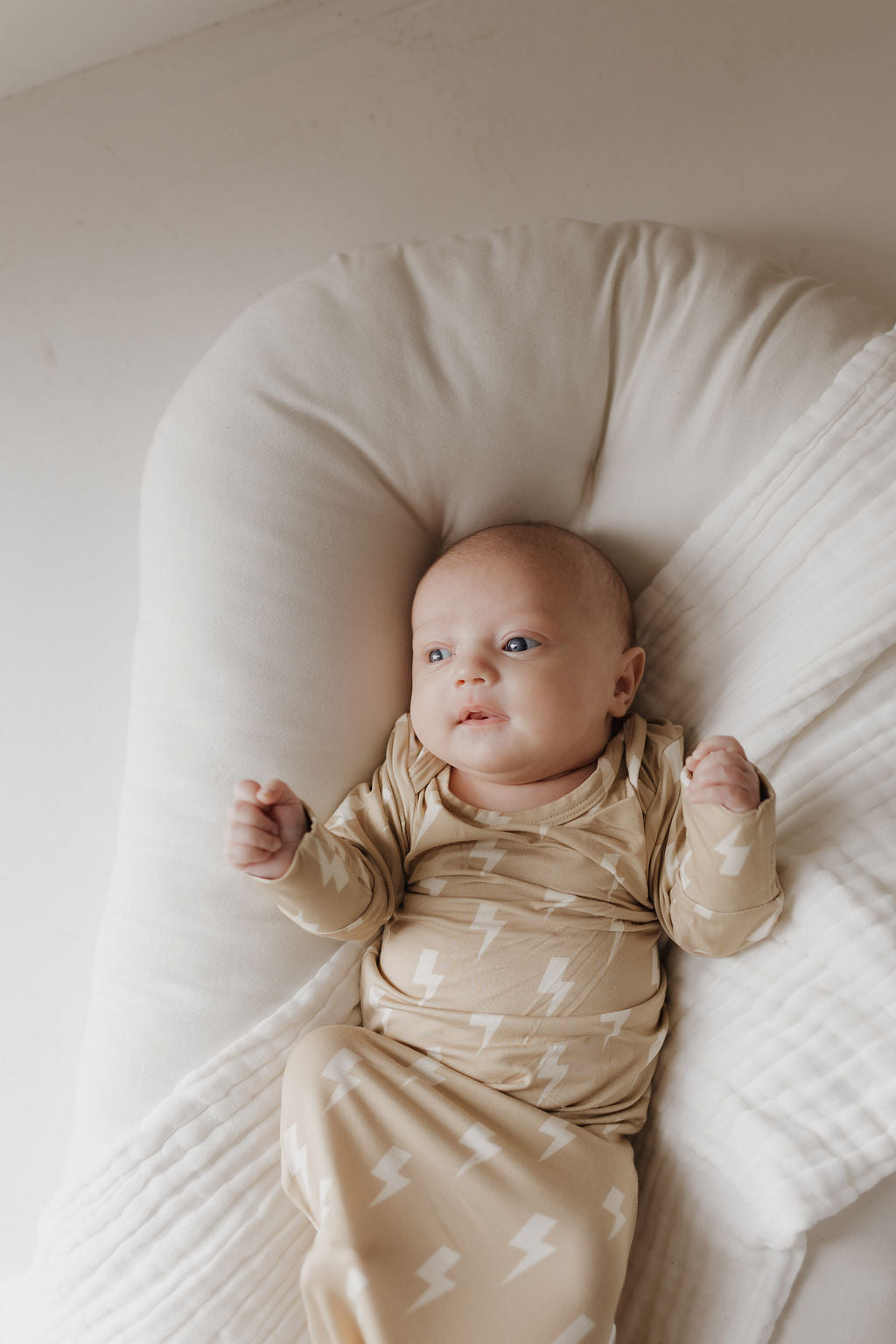 In a bright, serene setting, an infant gazes upward while resting on a soft, hypoallergenic cushion. They are wrapped in a bamboo blanket adorned with white lightning bolt patterns and wear the matching Bamboo Knotted Gown in Tan Lightning Bolt by forever french baby.