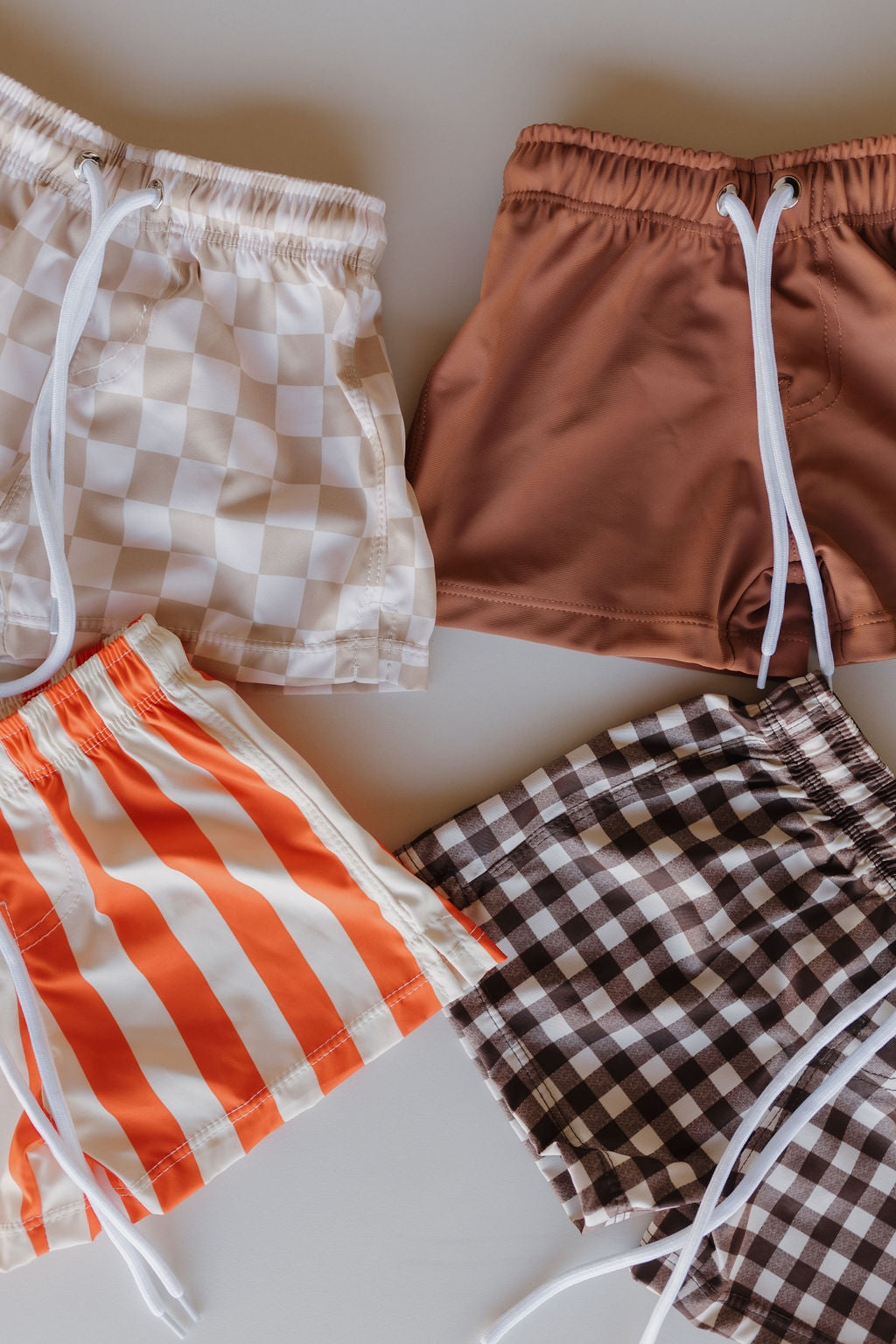 Four pairs of Child Boardshorts from the Terra collection by forever french baby are displayed on a light surface. Featuring white drawstrings and customizable fits, the designs include checkered white, solid brown, orange and white striped, and black and white checkered styles.