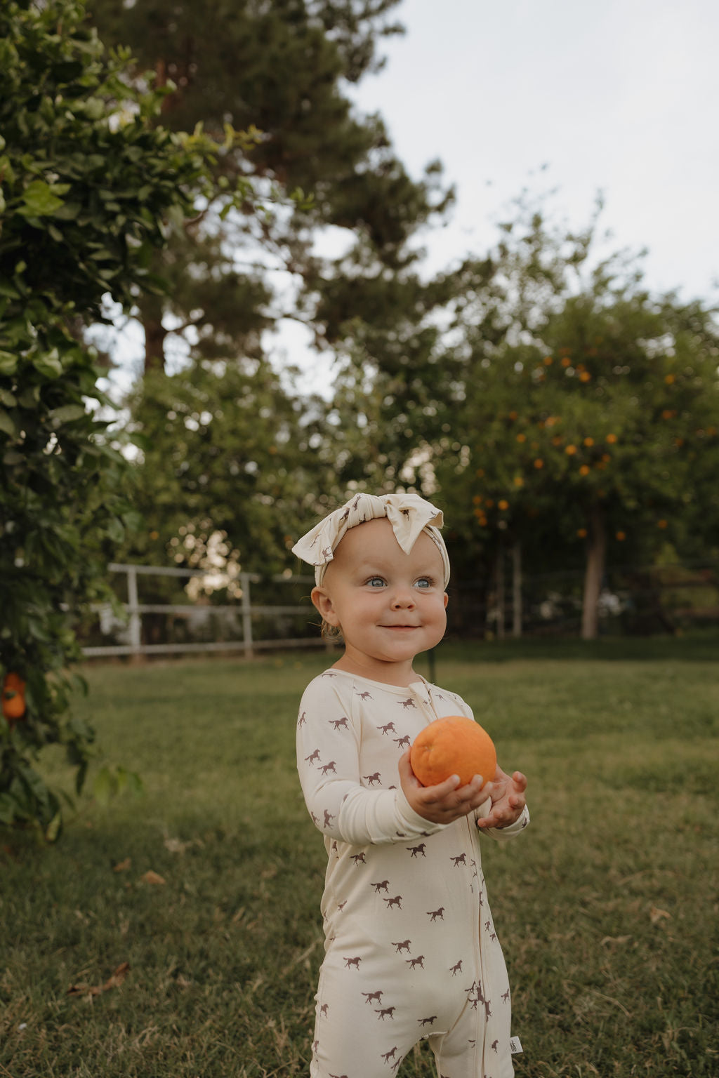 A smiling baby stands on grass in a garden, holding an orange. She wears a forever french baby onesie with animal prints and the Bamboo Head Wrap | Wild Horses by forever french baby, with green trees in the background.