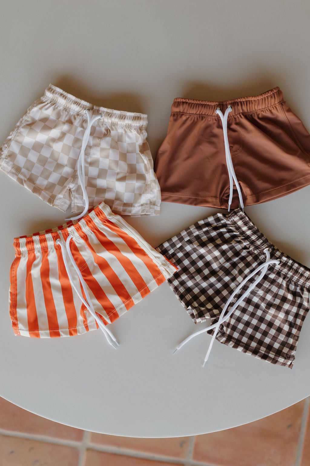 Four "Child Boardshort | Terra" pairs from forever french baby are arranged in a circle: clockwise from top left, beige checkered, solid brown, brown checkered, and orange striped. Each softens fit with a white drawstring.