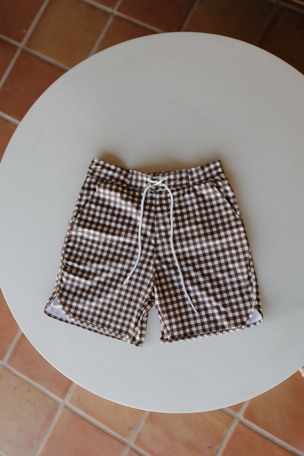 From the "forever french baby" Chocolate Gingham collection, these men's boardshorts blend brown and white checks with a stylish white drawstring. Displayed flatly on a round white table against a tiled floor, they offer a captivating twist on classic men's beachwear.