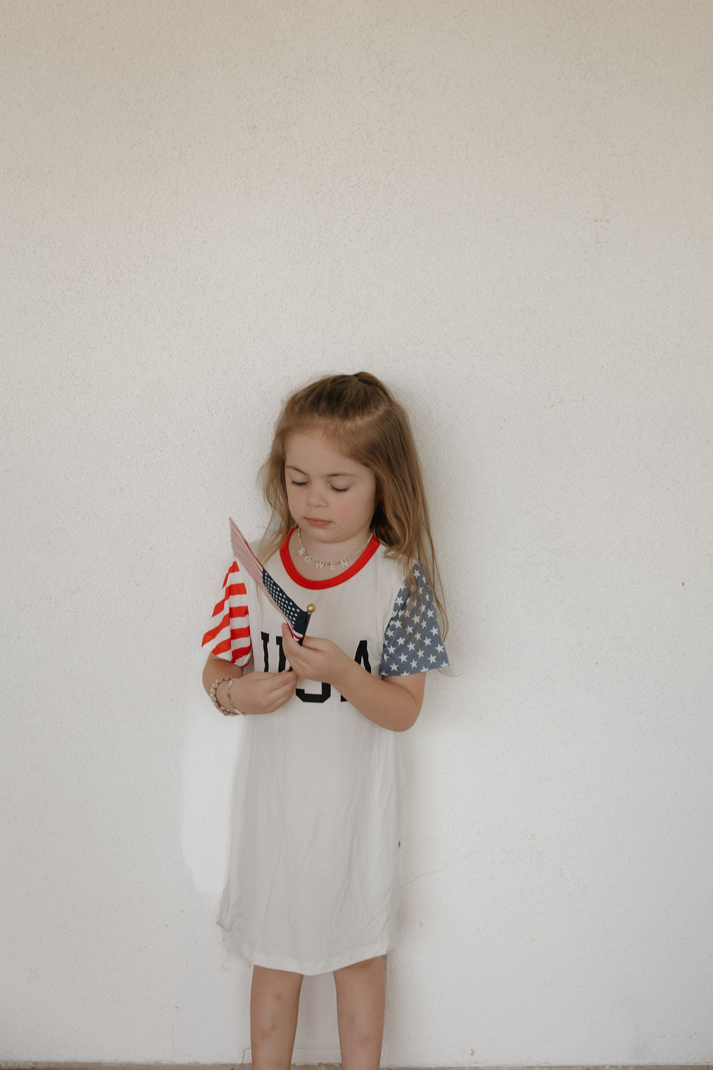 A young girl stands against a white wall, wearing the forever french baby Bamboo T-Shirt Dress | Stars & Stripes with "USA" print and patriotic sleeves, while looking down and holding a small American flag.