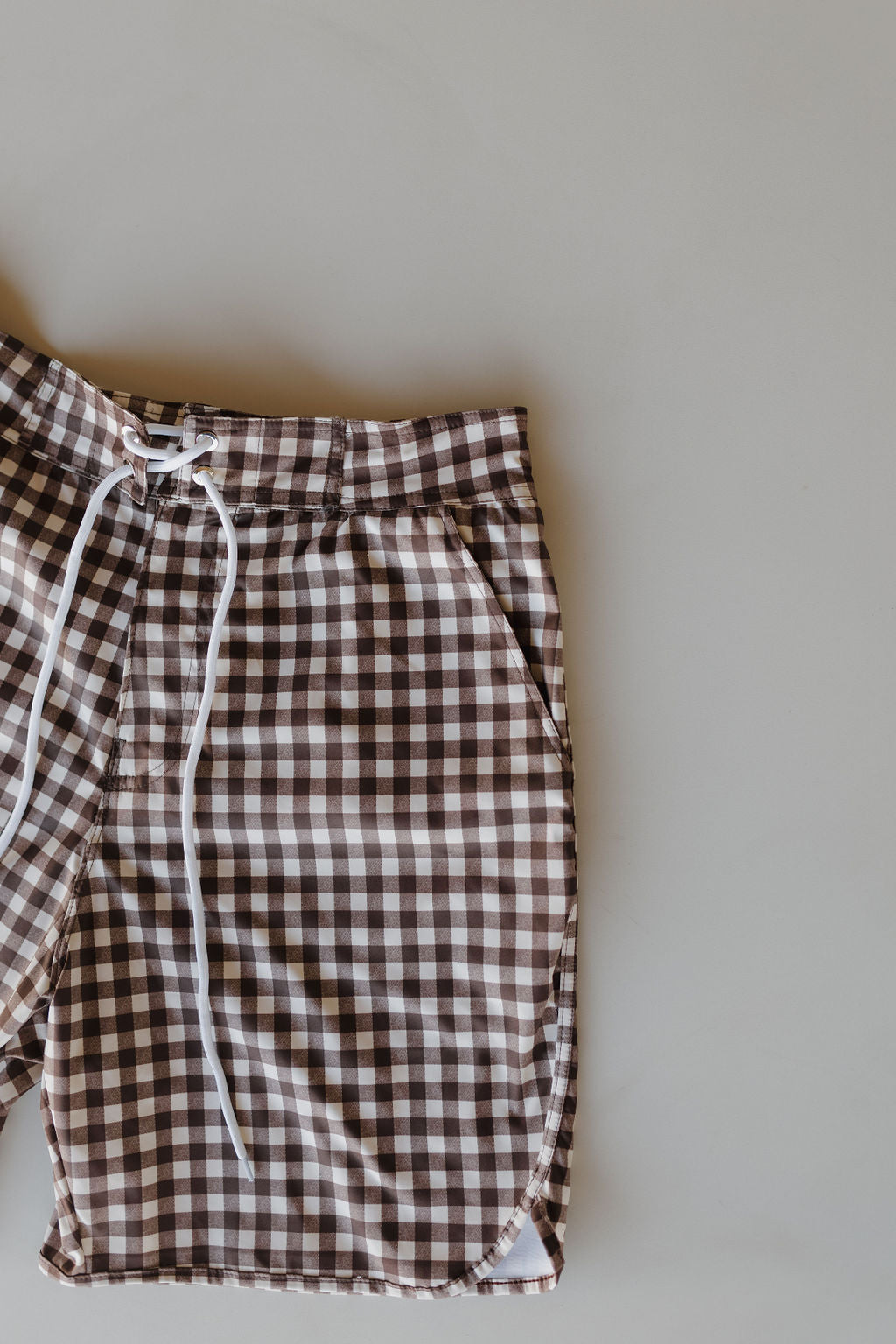 From the forever french baby collection, the Men's Boardshort in Chocolate Gingham flaunts a brown and white checkered pattern with a white drawstring waistband. The image stylishly showcases half of these trendy shorts against a neutral backdrop.