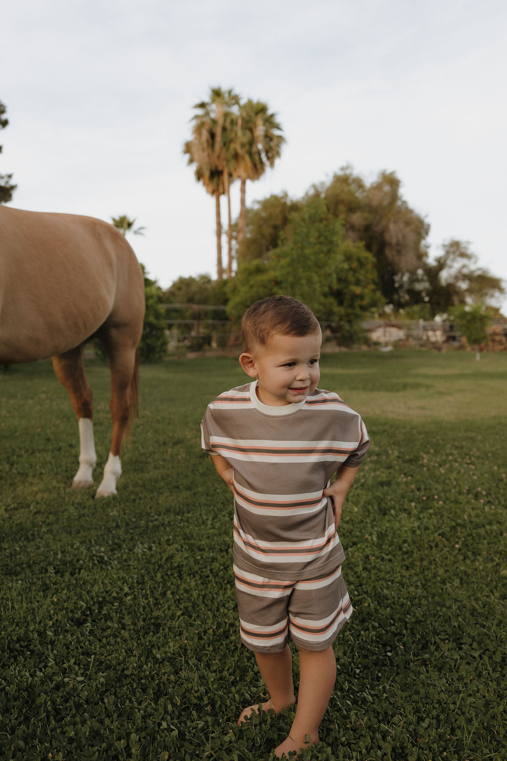 A young boy wears the forever french baby Child Short Set | Sedona Stripe, standing barefoot on grass with his hands behind his back, a horse beside him and trees in the background.